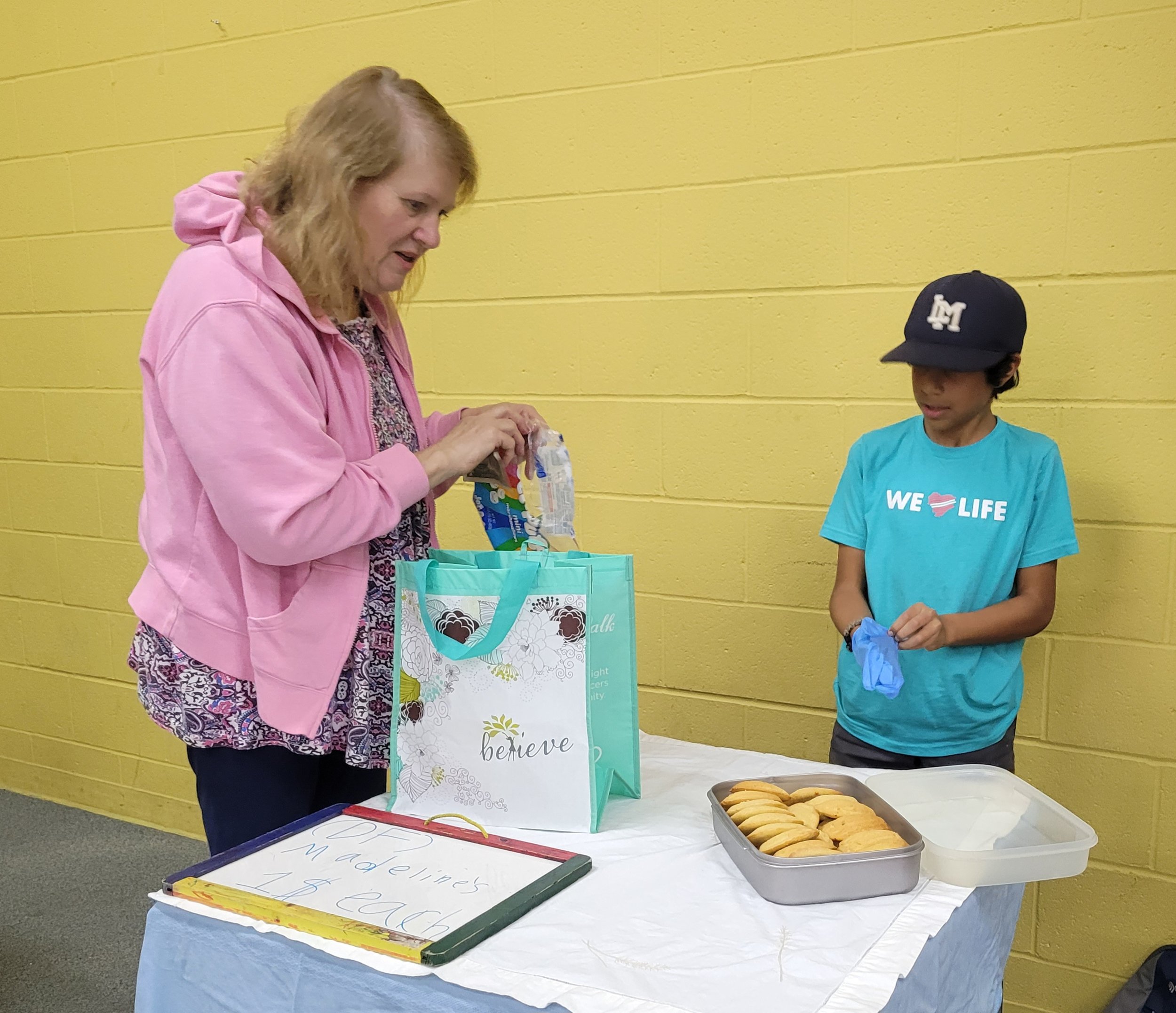 A woman and a boy standing at a table with cookies in trays and a gift bag, against a yellow wall.