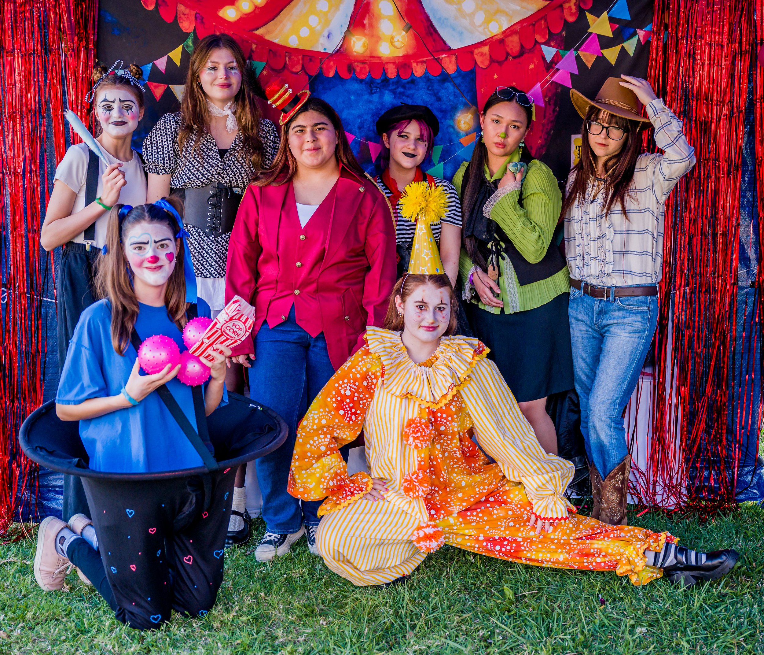 Group of children dressed as clowns and circus performers at a festive event, standing in front of a colorful circus-themed backdrop with red, blue, and yellow decorations.