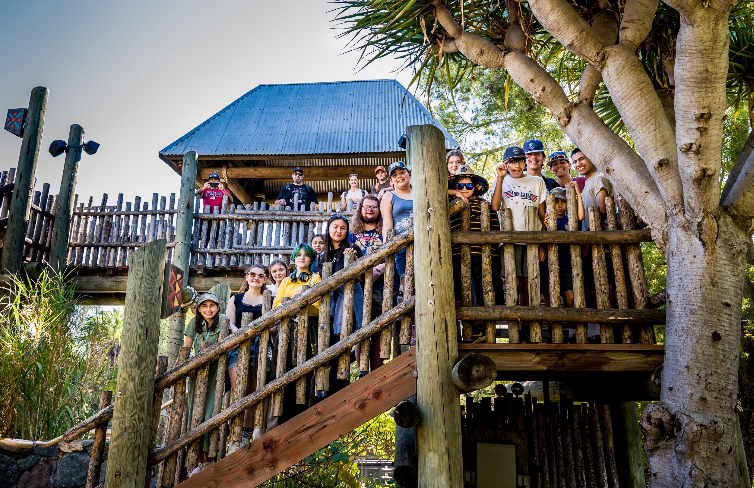 A group of children and adults standing on a wooden balcony by a tree, smiling and posing for a photo on a sunny day.