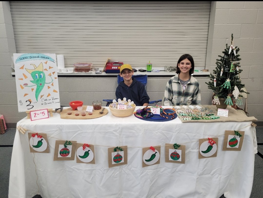 Two children sitting behind a table at a holiday market stand, decorated with handmade Christmas ornaments, a small decorated Christmas tree, and a festive banner. The boy is wearing a yellow cap and navy hoodie, and the girl is in a plaid shirt. The stand features a sign with a green chili pepper character and sells salsa for $3.