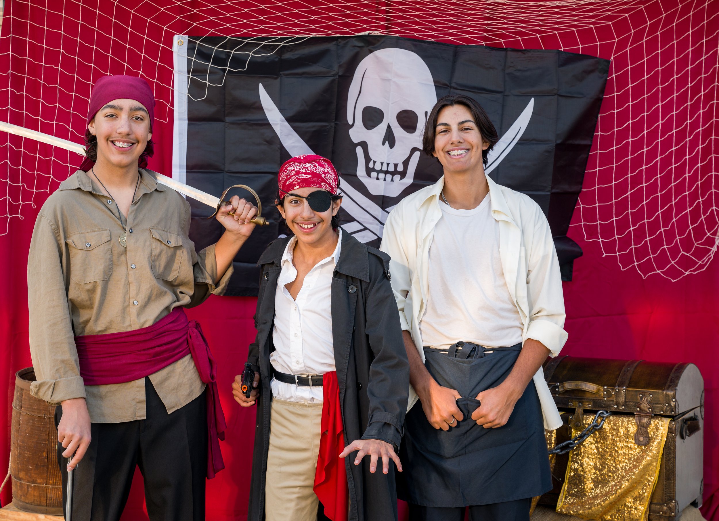 Three young people dressed as pirates standing in front of a pirate flag with a skull and crossbones symbol, inside a red tent with netting.
