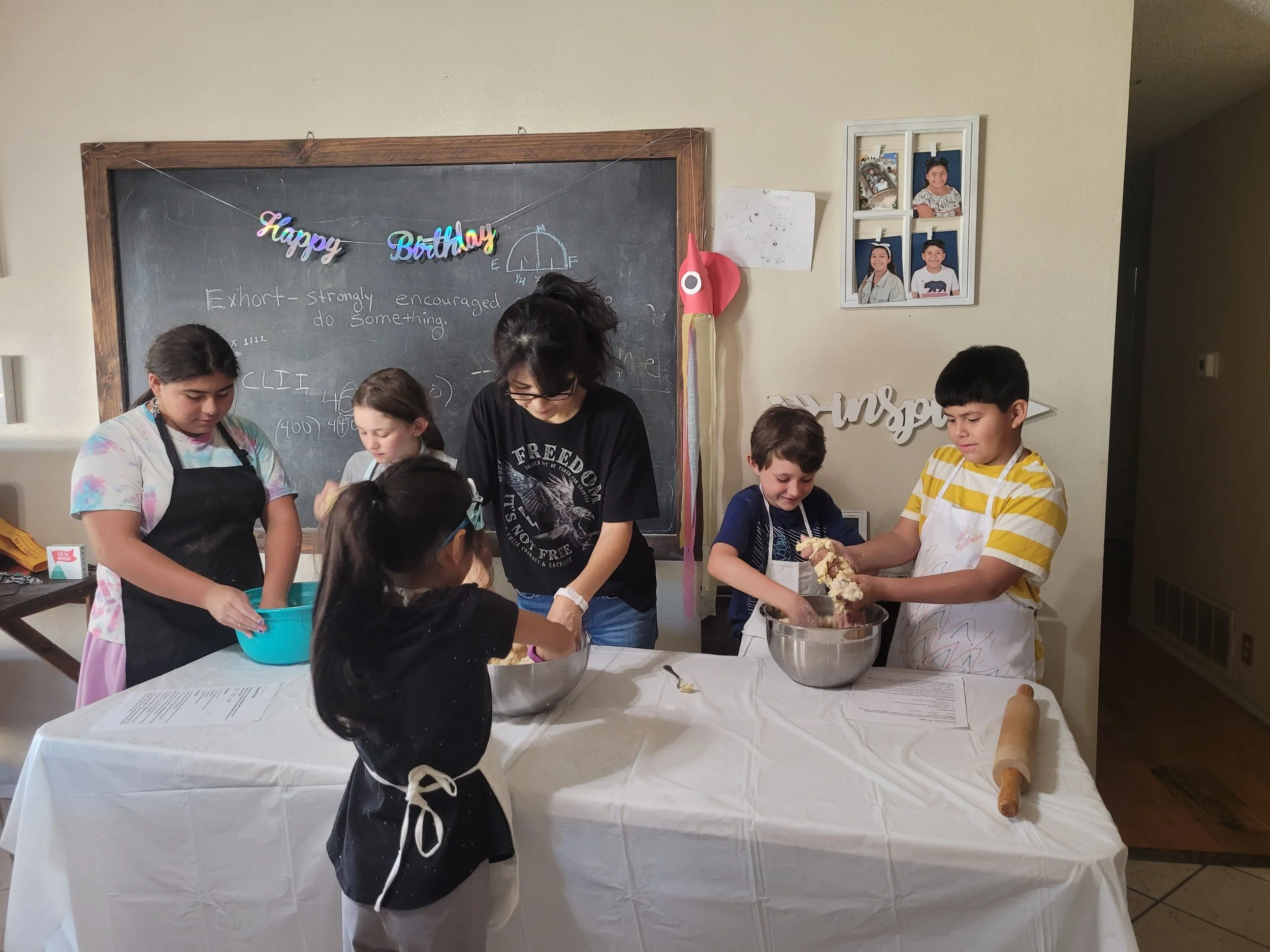 Children and a woman baking or cooking together at a kitchen table, with bowls, a rolling pin, and papers.