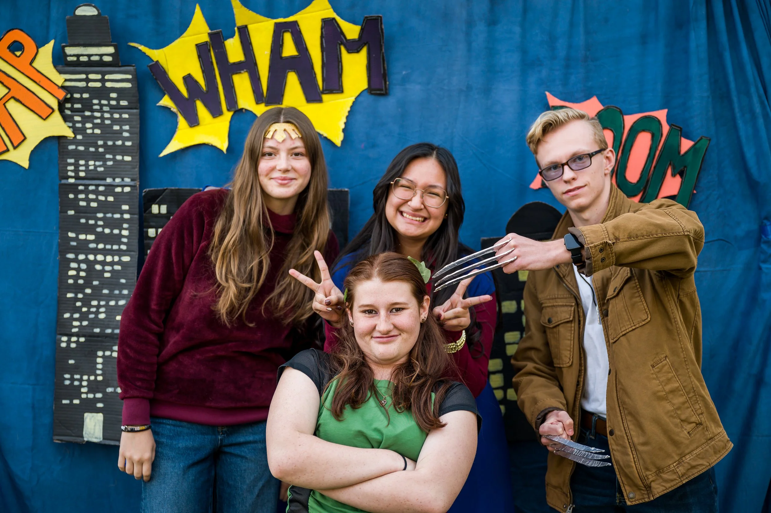Group of five young people posing together in front of a blue background decorated with comic book style signs and cityscape illustrations, smiling and making playful gestures.