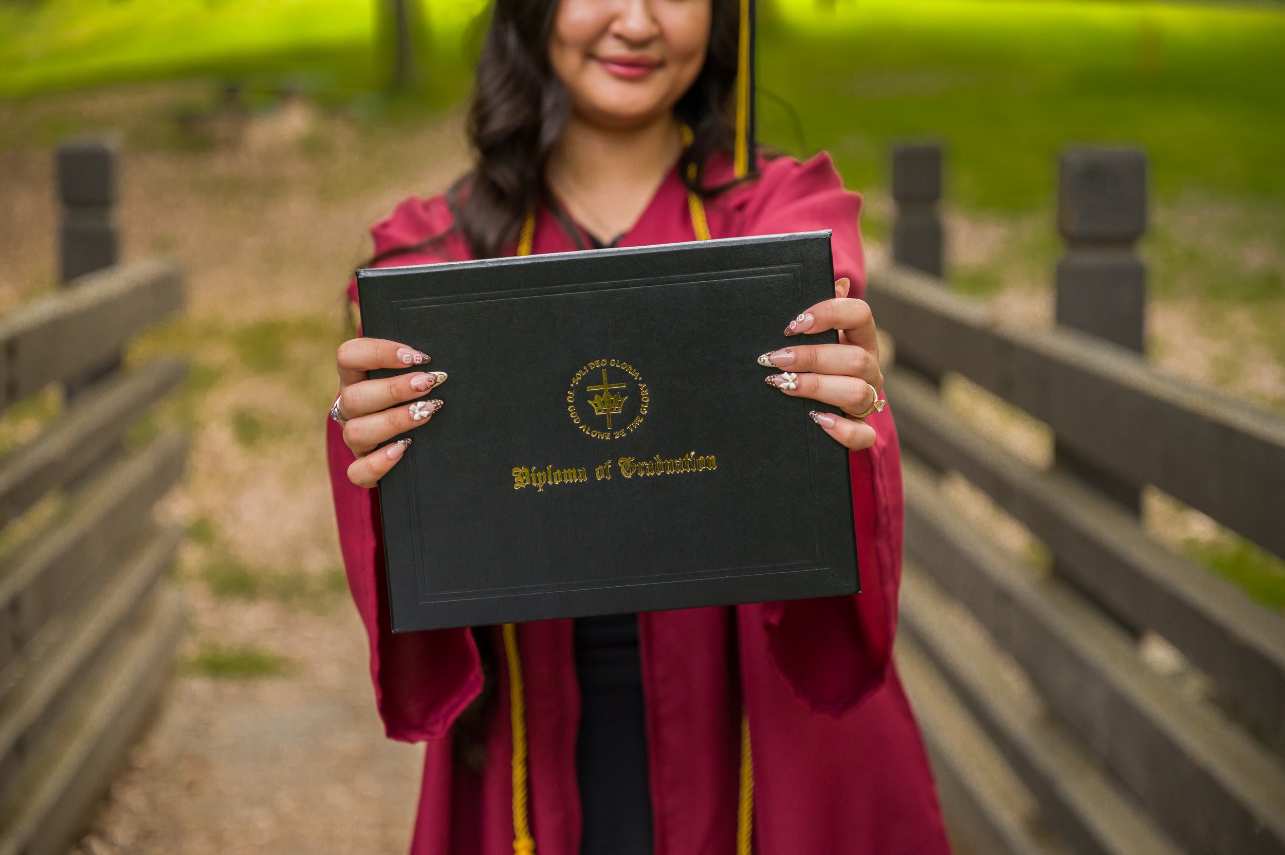 Person holding a diploma of graduation with a smile, standing outdoors on a wooden bridge.