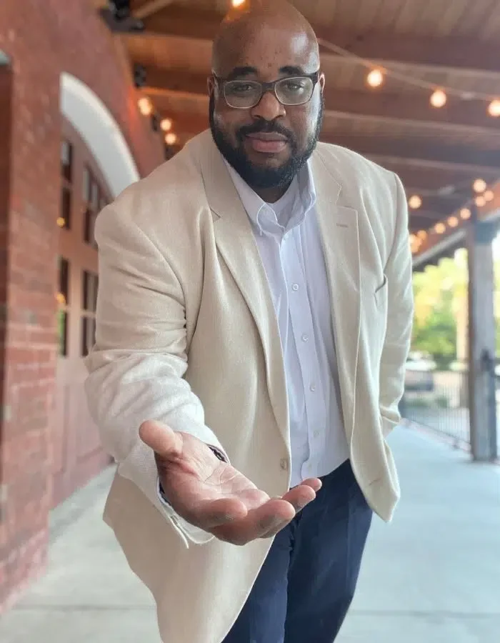 Man with glasses in a light-colored blazer and white shirt extending his hand forward in an inviting gesture outdoors.