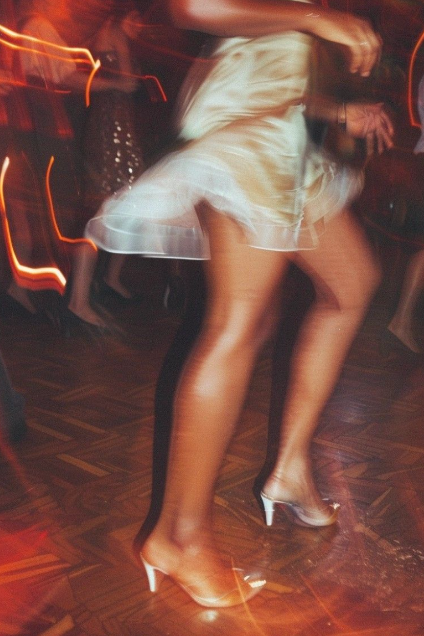 A person dancing in a white dress and high heels at a party or nightclub, with blurred motion and colorful lights in the background.