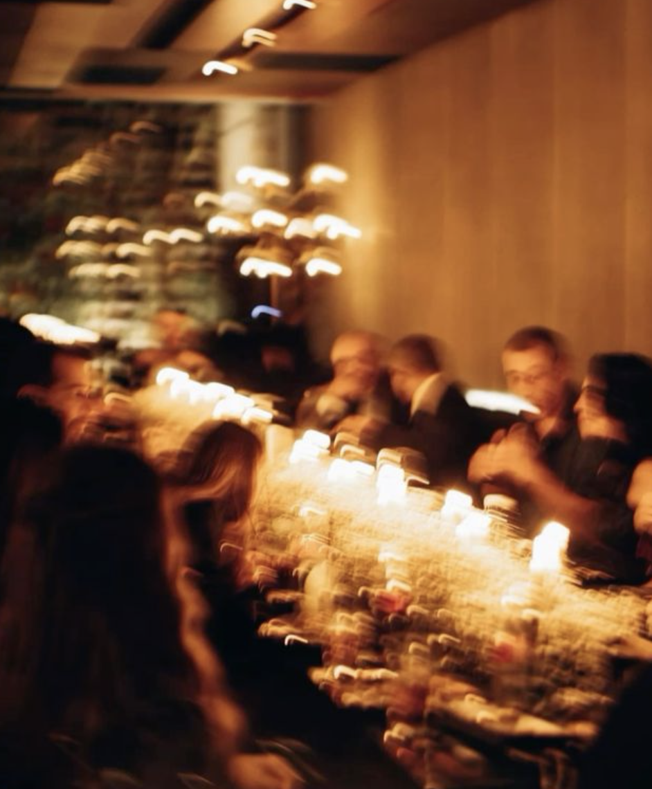 Blurred image of people sitting at a long table with candles, in a dimly lit restaurant or event space.