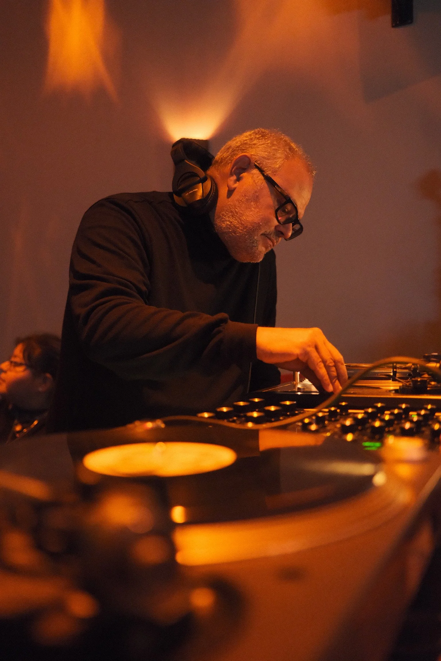 A male DJ wearing glasses and headphones, focused while operating a DJ mixing console in a dimly lit setting with orange lighting.