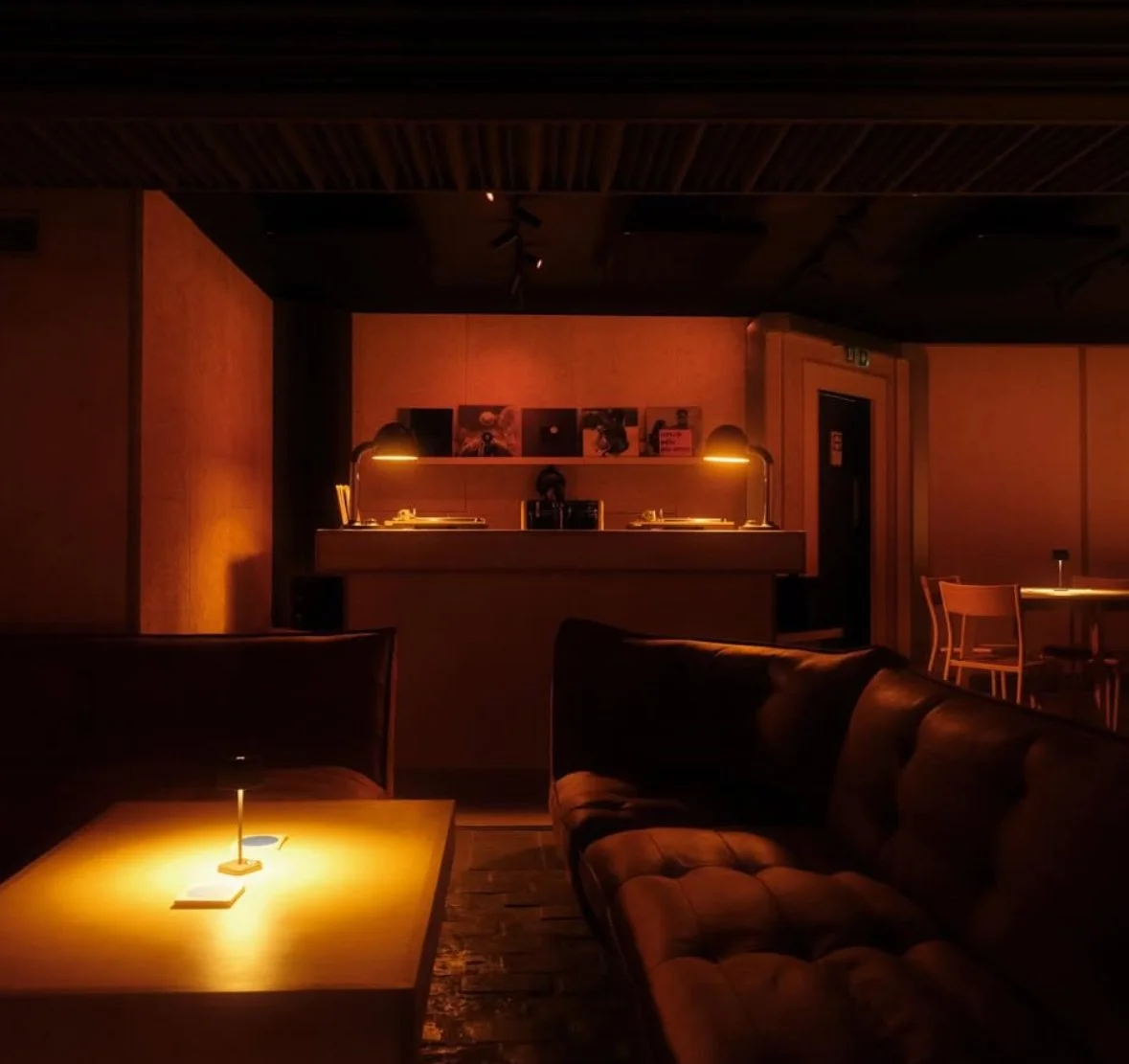 Dimly lit lounge area with a coffee table, a sofa, and a bar in the background illuminated by warm yellow lighting.
