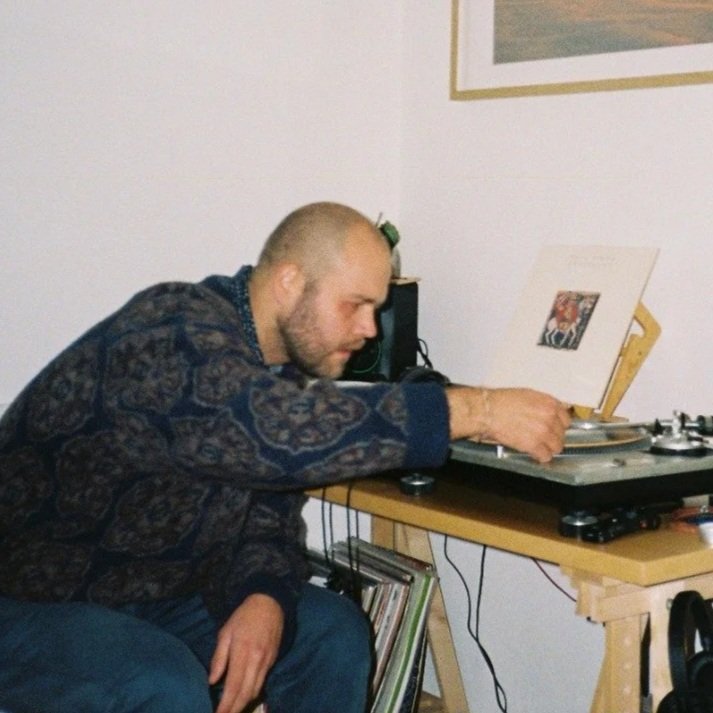 A man with a beard and short hair, wearing a patterned sweater, is sitting at a desk adjusting a record player.