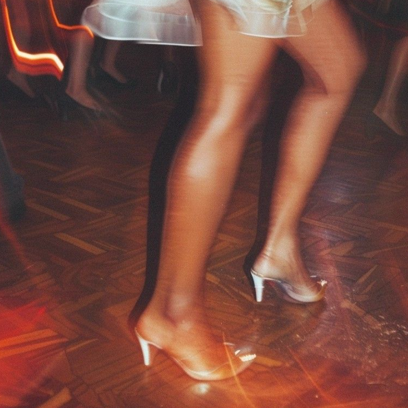 The image shows a person wearing high-heeled shoes and sheer stockings, sitting at a table on a wooden floor, with neon lights visible in the background.