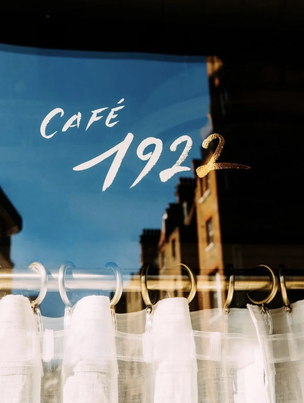 A reflection in a café window showing the text 'Café 1922' and the number 2 in gold, with buildings and a blue sky in the background, while white curtains hang below.