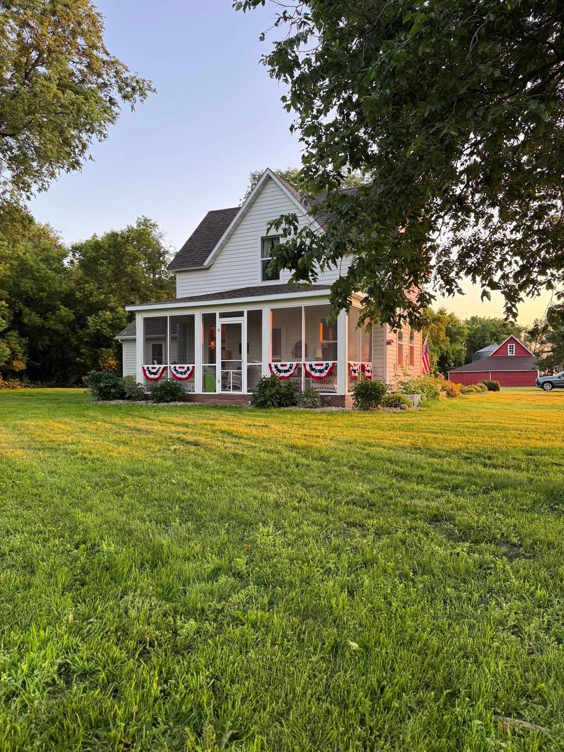 A white two-story house with a screened porch decorated with patriotic red, white, and blue bunting, set on a lush green lawn during sunset.