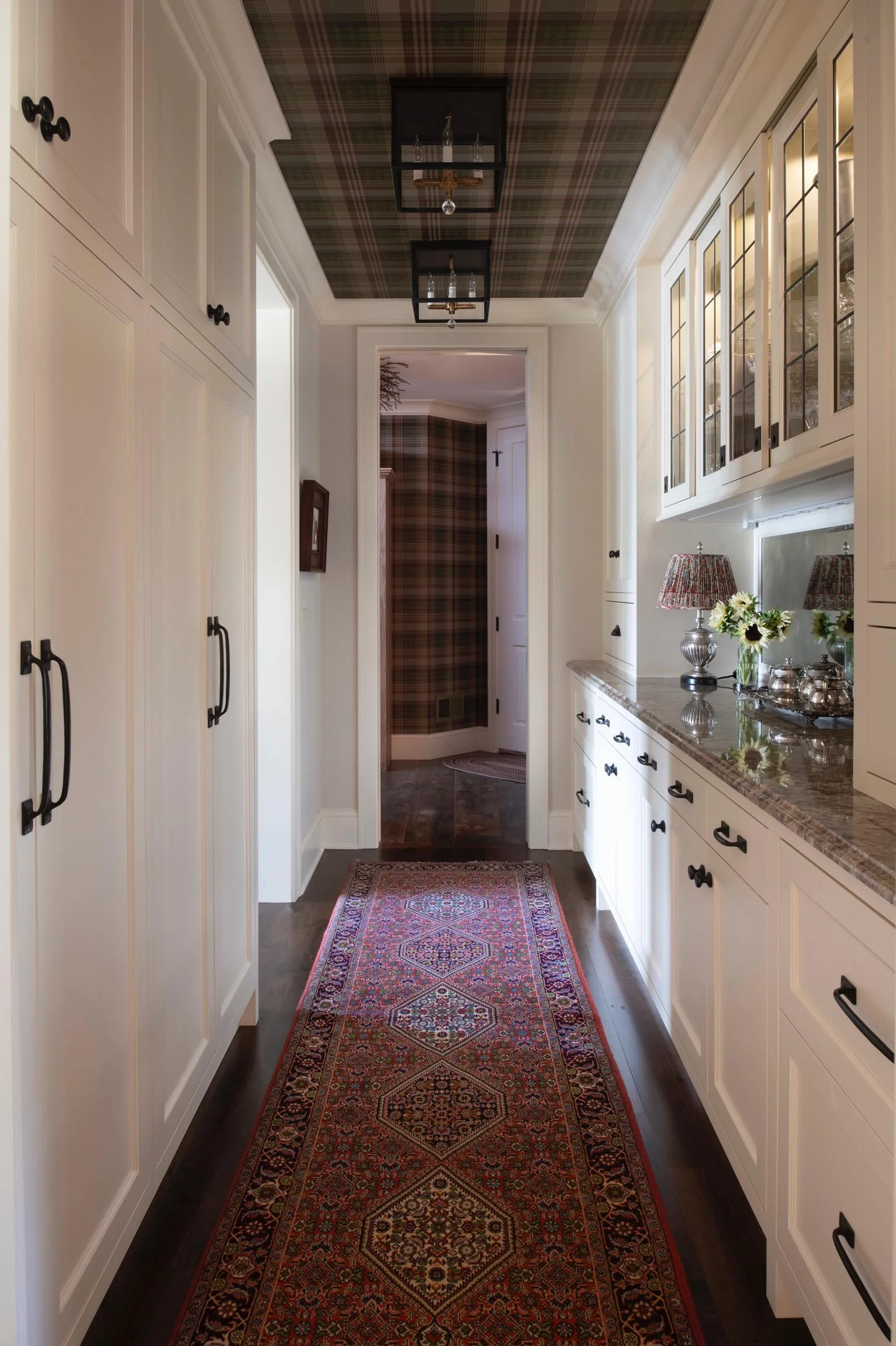 A hallway with white cabinets on both sides, dark hardwood flooring, a colorful patterned rug, and a view into a room with plaid wallpaper. There's a window with glass-paneled cabinets above, and decorative lamps with floral arrangements on a countertop.