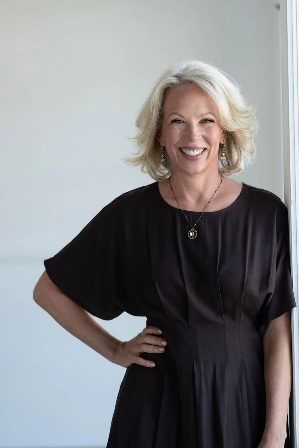 A smiling middle-aged woman with blonde hair wearing a black dress and jewelry, standing by a window.