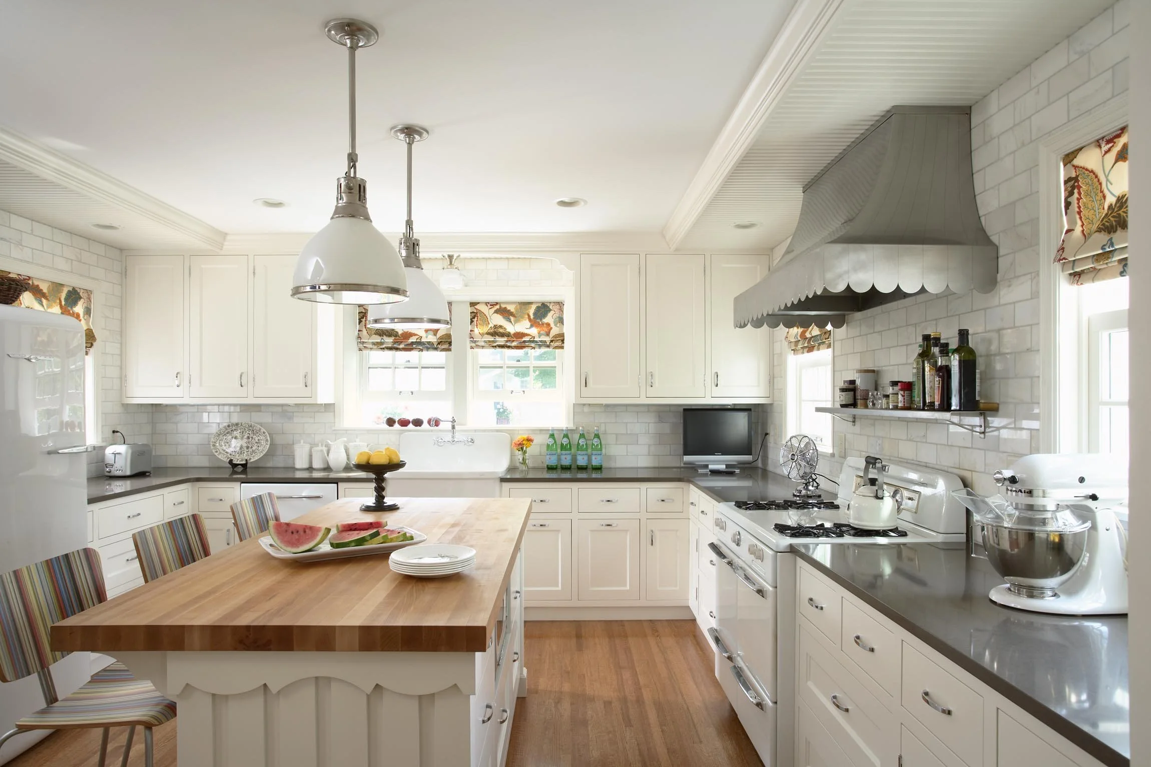 A bright kitchen with white cabinets, a wooden island with striped chairs, hanging white pendant lights, a white stove, and a window with colorful floral curtains, featuring a sink under the window and a small TV on the counter.