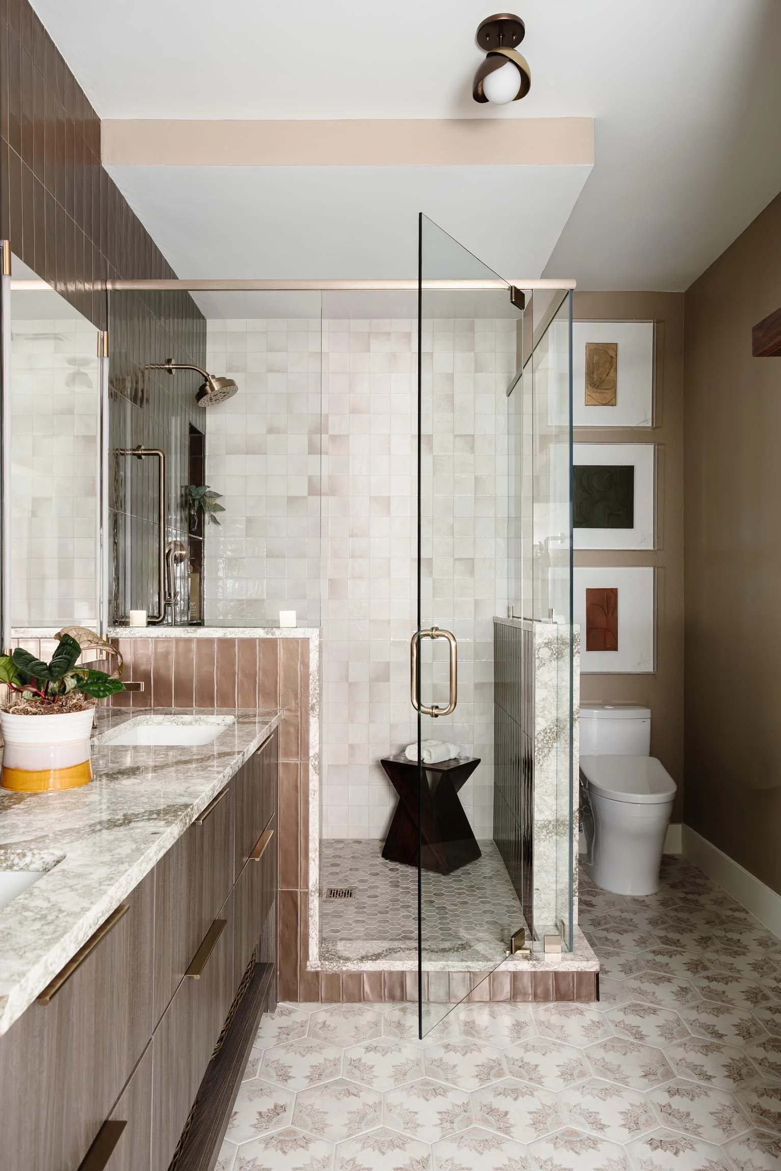 A modern bathroom with a glass shower enclosure, beige and brown tiles, a dual vanity with a marble countertop, potted plant, and decorative wall art.