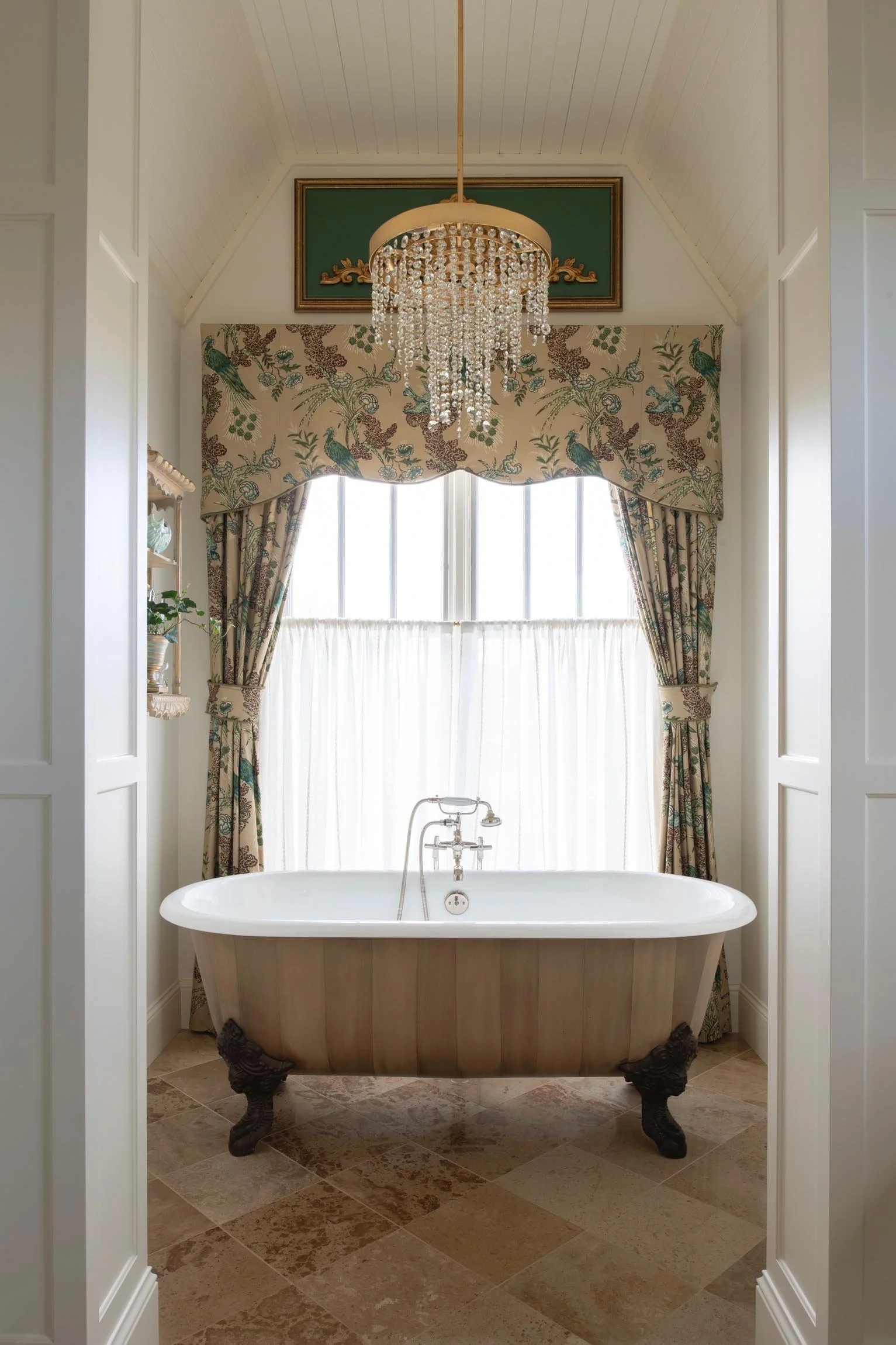 A vintage bathtub with clawfoot design in a bathroom with cream-colored walls, a chandelier, and a window with floral curtains.
