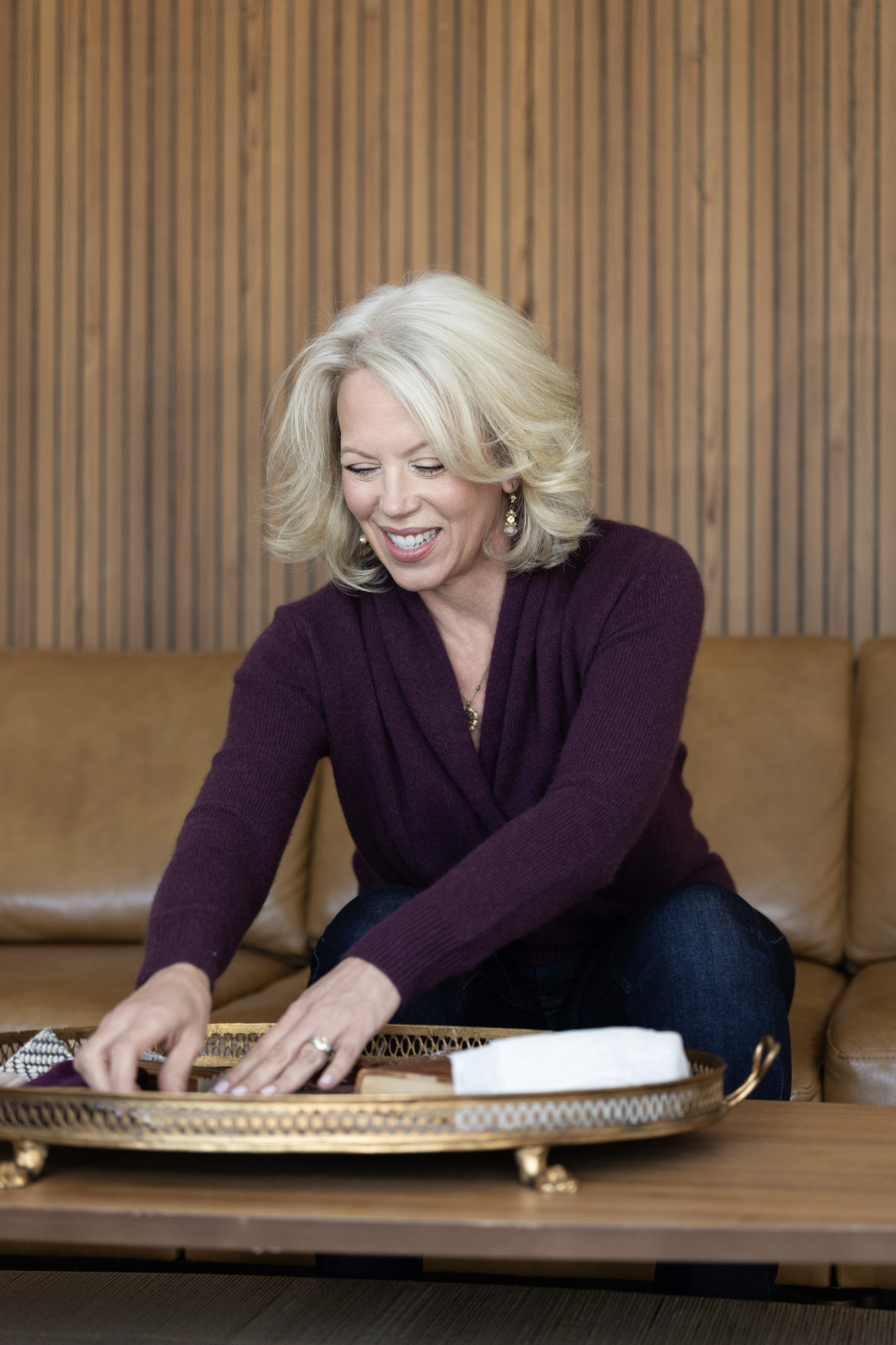 A woman with blonde hair, wearing a purple sweater, smiling and reaching into a decorative tray on a wooden table, sitting on a tan sofa with a wood-paneled wall background.