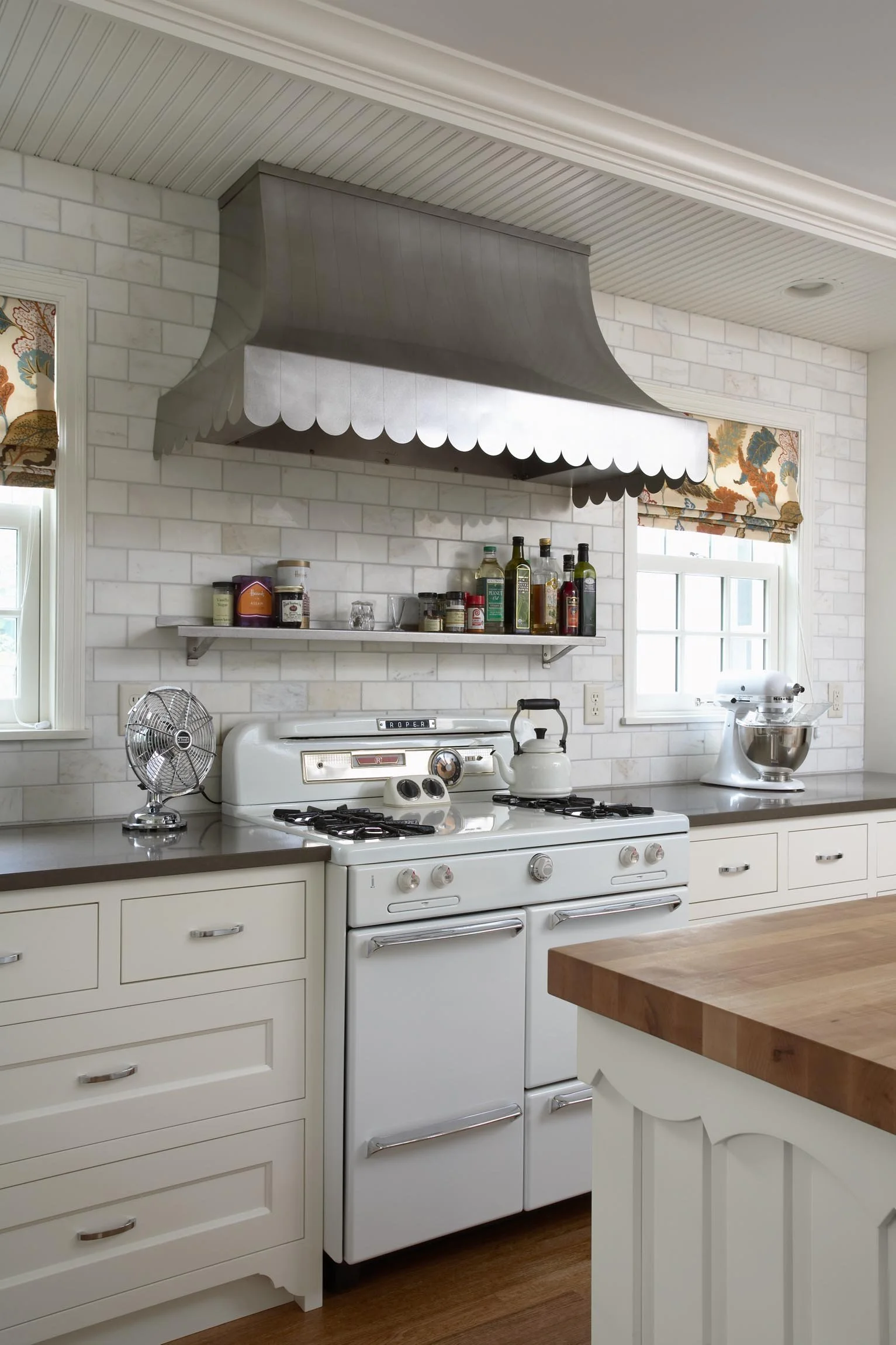 A kitchen with white cabinets, a white gas stove, a wooden countertop, a metal range hood, and a shelf with spices and oils. There is a window with floral curtains and a small fan on the counter.