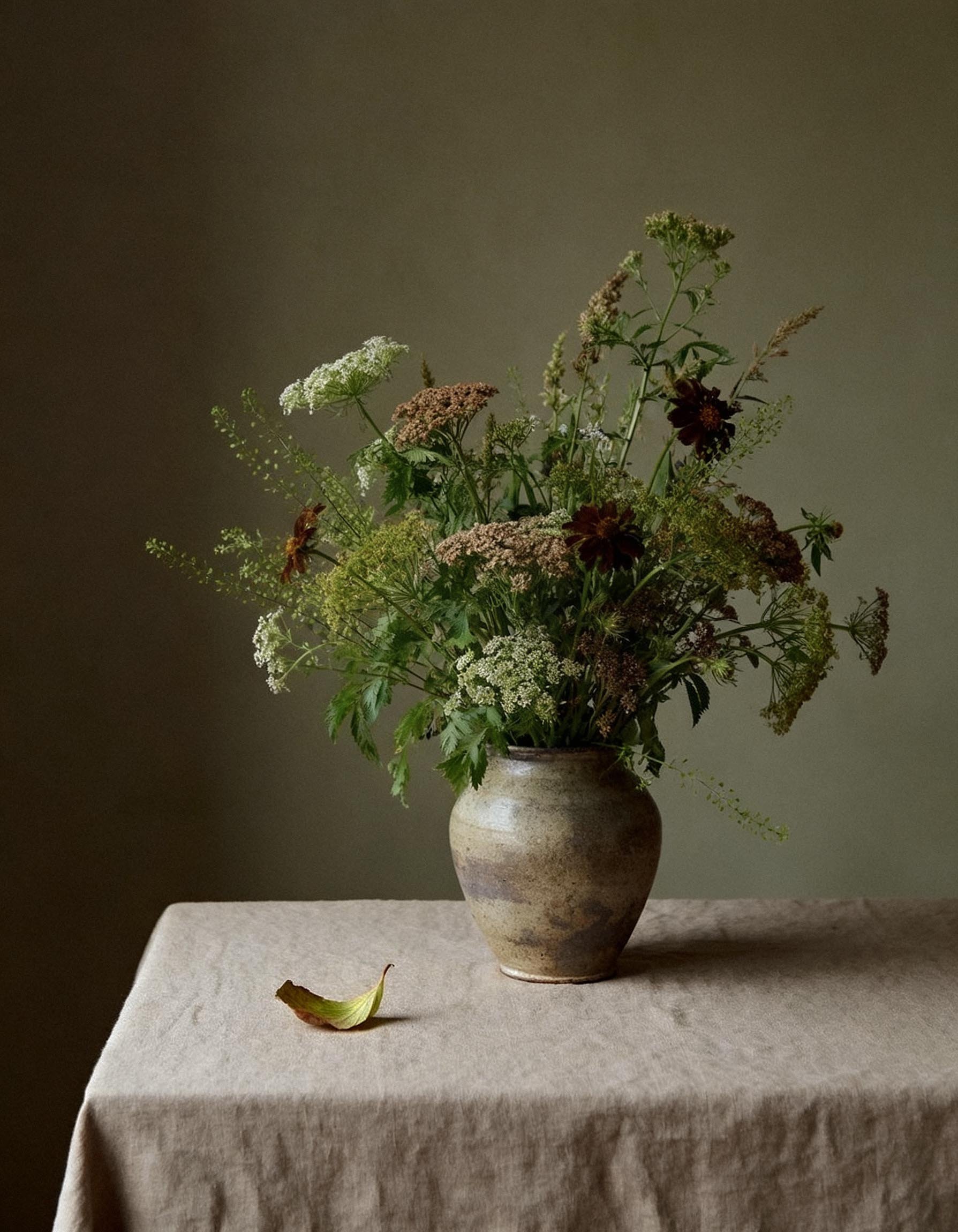 A ceramic vase filled with various wildflowers and greenery, placed on a textured fabric-covered table with a neutral-colored background.