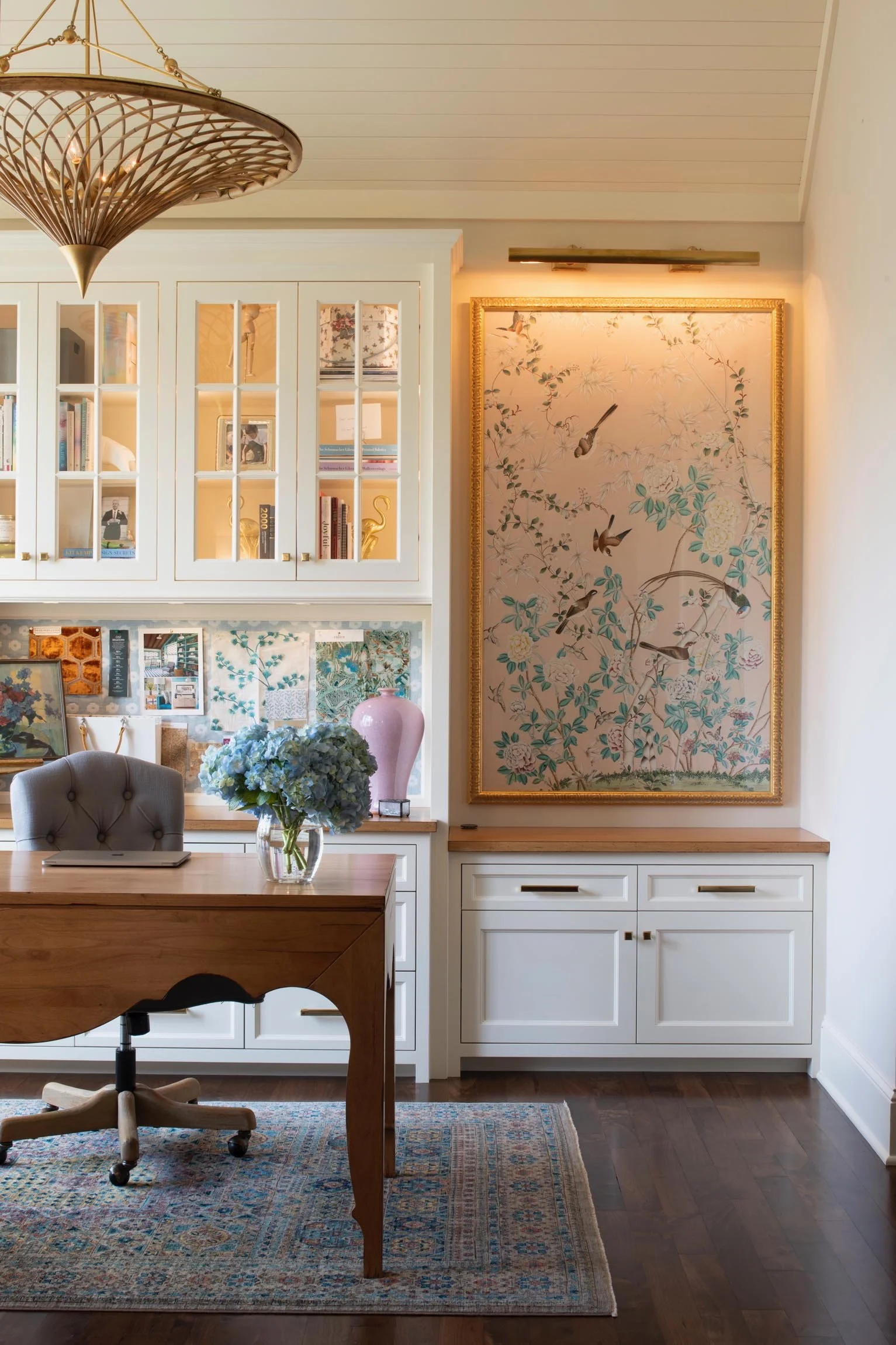 A home office or dining area with a wooden table, a gray swivel chair, a floral arrangement in a glass vase, built-in white cabinetry with glass doors displaying books and decorative items, a large framed floral and bird painting, a colorful patterned rug, a pink vase, and a light fixture on the ceiling.