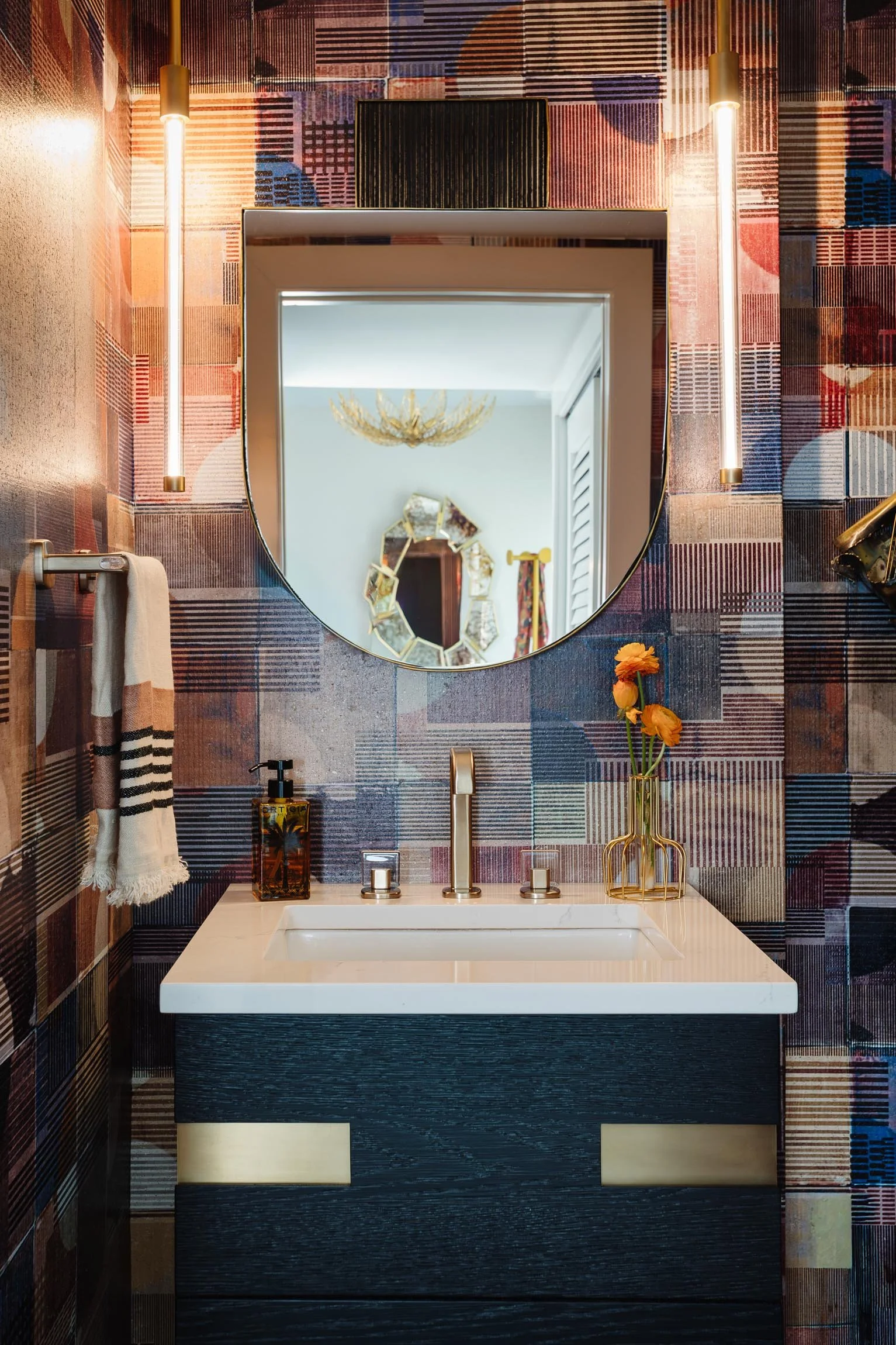 A bathroom with a black vanity, white sink, gold fixtures, and a mirror reflecting a chandelier and part of a living space, with colorful, geometric-patterned tiles on the wall and a vase with orange flowers on the counter.