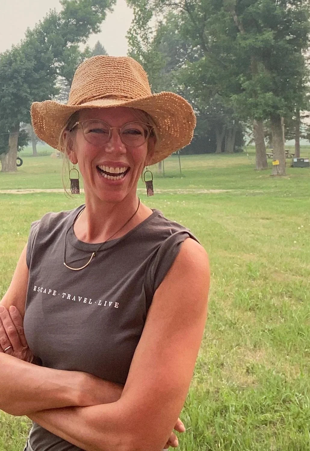 A woman smiling and standing outdoors in a park wearing a wide-brimmed straw hat, glasses, earrings, a gray T-shirt with white text, and a necklace.