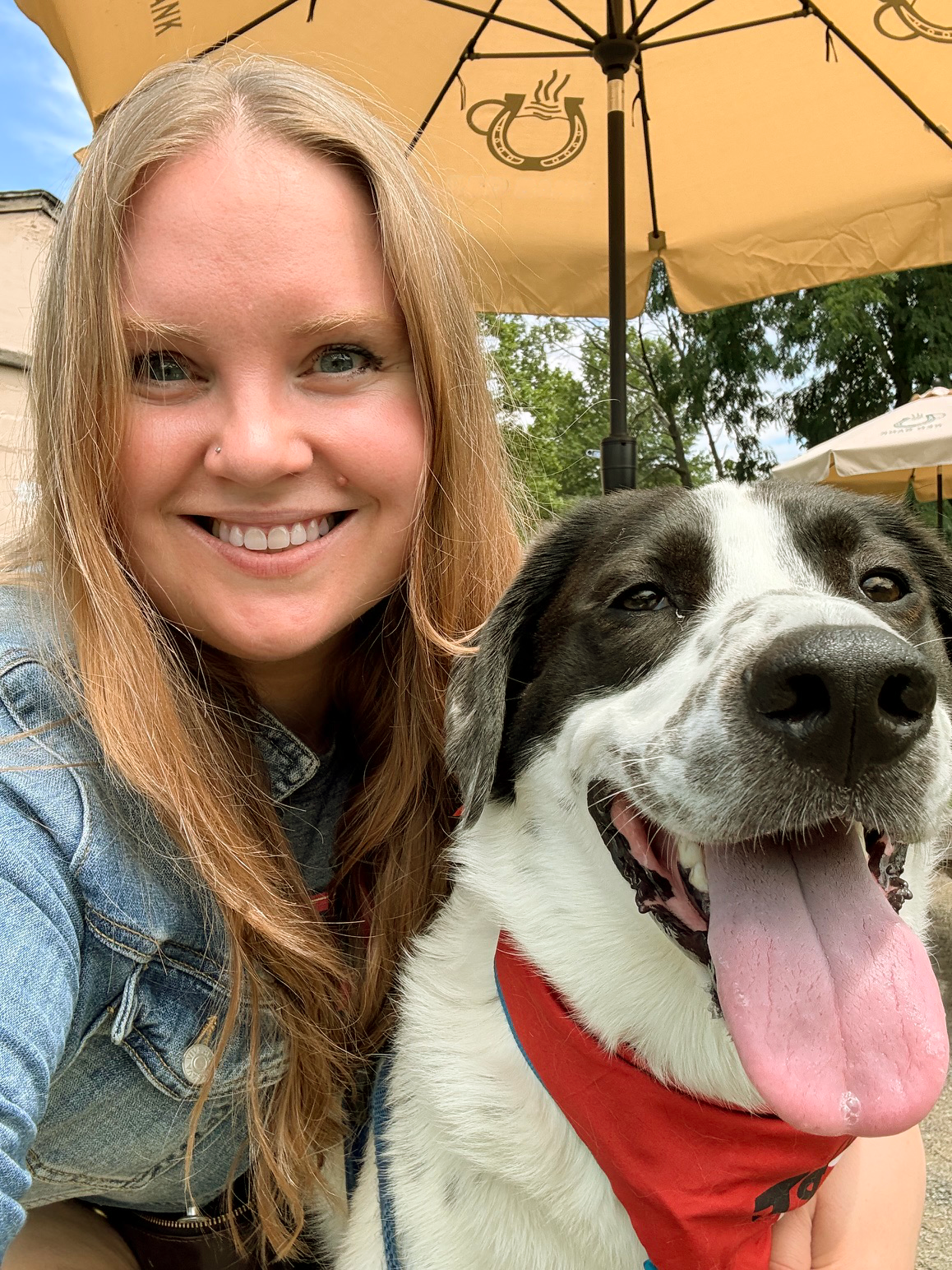 A smiling woman with long blonde hair and a nose piercing taking a selfie with a happy black and white dog wearing a red bandana outdoors under a yellow umbrella.