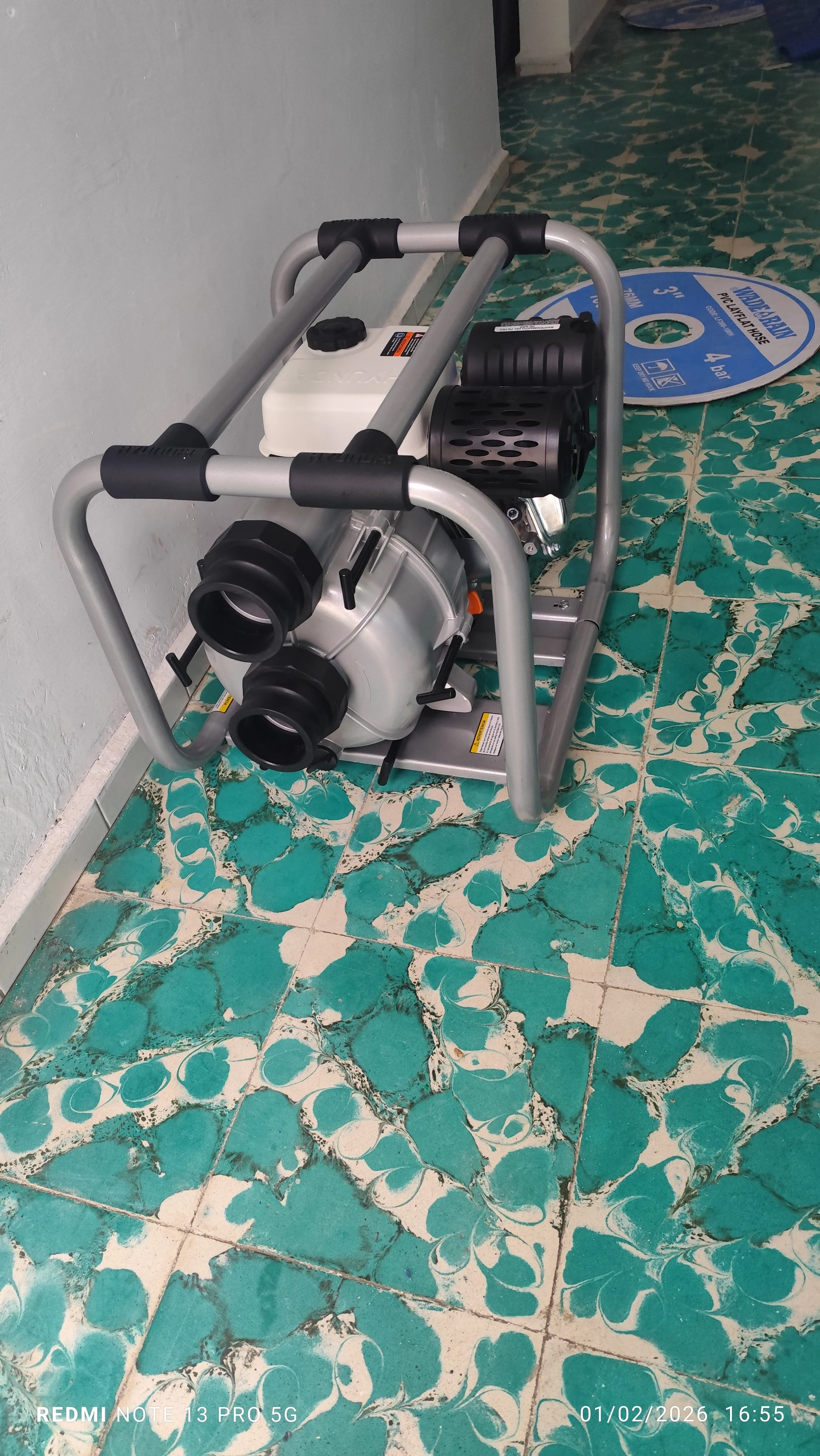 Equipamiento de limpieza, probablemente una hidrolavadora, colocado en el suelo junto a un rollo de malla para el lavado con manguera, en una habitación con pisos de azulejos verdes con patrones blancos y paredes grises.