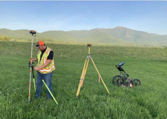 Persona realizando mediciones con equipo topográfico en un campo verde con montañas al fondo.
