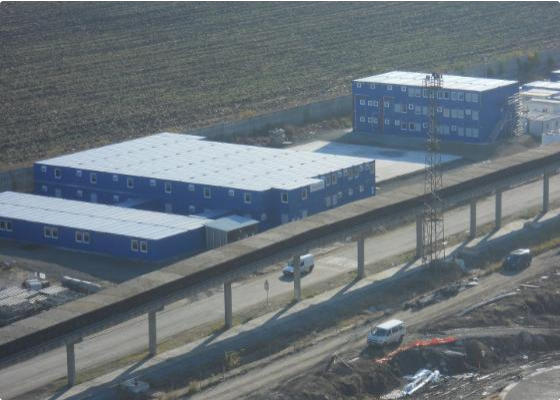 Edificios azules en un área industrial junto a una autopista elevadas, con vehículos circulando debajo y campo abierto en el fondo.