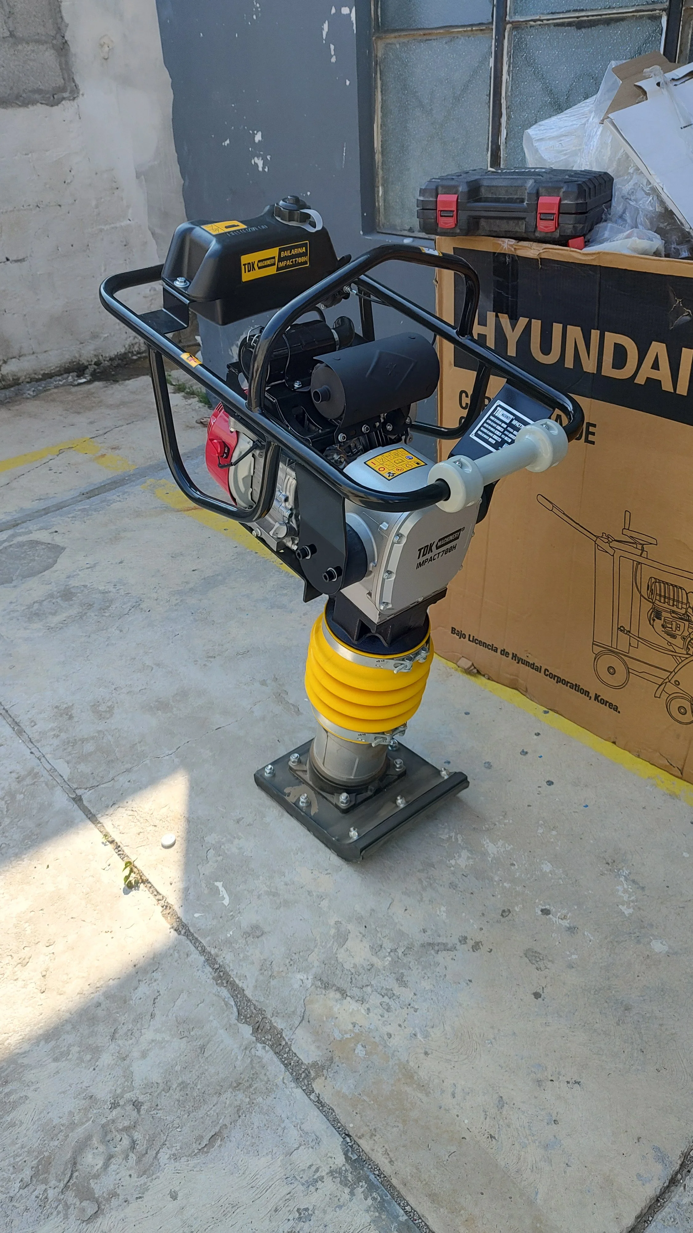 Taladro neumático con motor en la parte superior conectado a una base de hormigón, con una caja de herramientas y una caja de cartón con la palabra Hyundai en fondo.