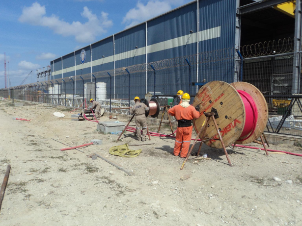 Cuatro trabajadores en ropa de seguridad y cascos trabajando en una obra en construcción en un sitio industrial, con una cerca de alambre y una estructura grande azul al fondo.