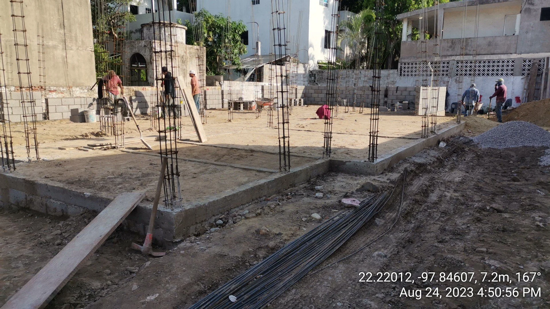Sitio de construcción con columnas de acero y encofrado, trabajadores en la obra, paredes en proceso, tierra y material de construcción en el terreno.