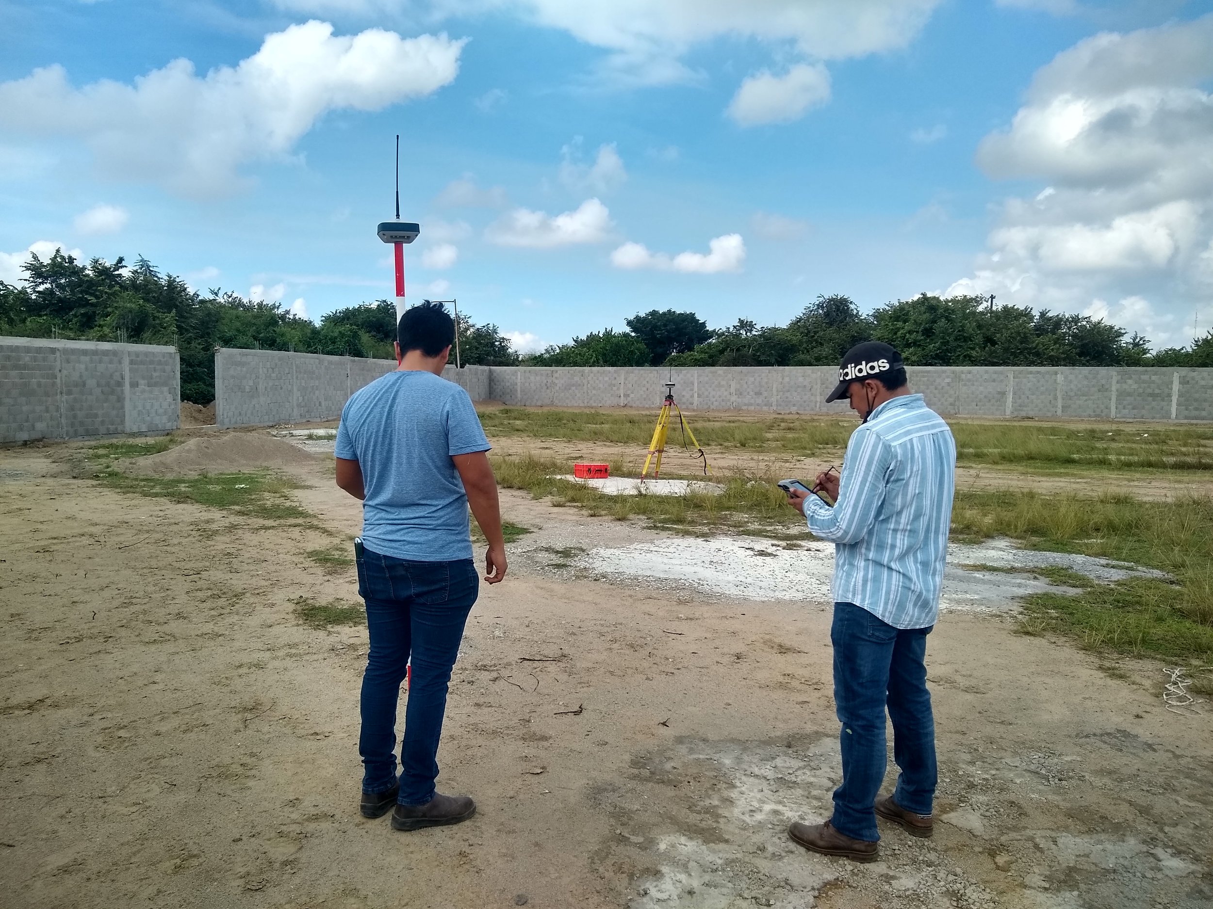 Dos hombres en un sitio de construcción observando un equipo de medición y una estación total montada en trípode. Uno de ellos lleva una gorra y el otro un teléfono móvil.