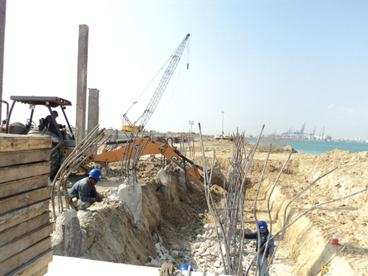 Construcción en curso en un sitio junto al mar, con maquinaria y trabajadores con cascos protegiendo cables y estructuras de acero.