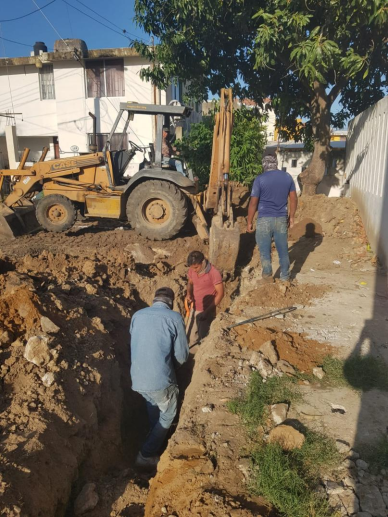 Tres personas trabajando en una excavación con maquinaria pesada en un entorno urbano, con árboles y casas en el fondo.