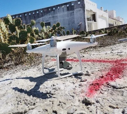 Un dron blanco en un terreno arenoso con cactus y un edificio blanco al fondo.