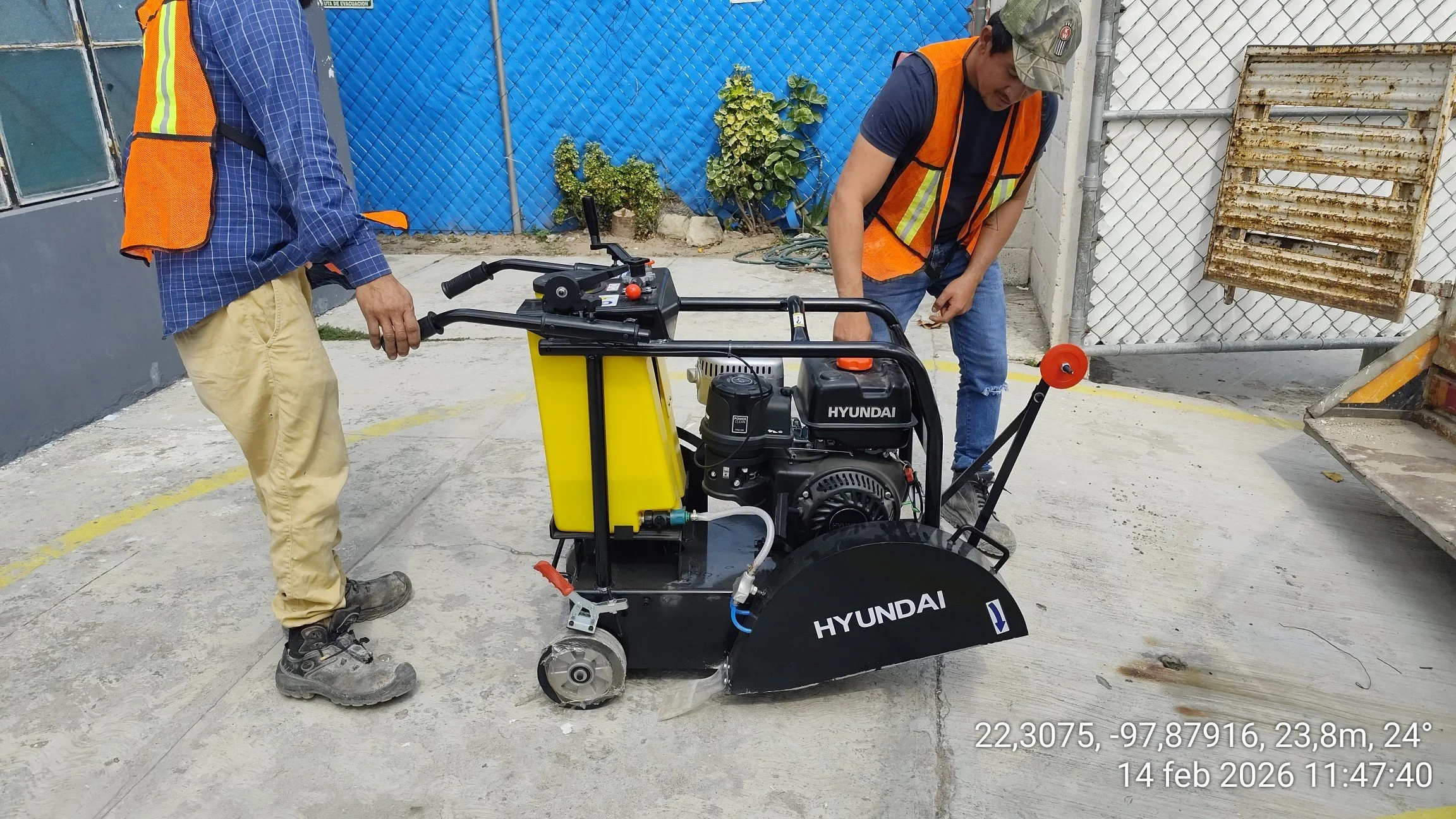 Dos trabajadores con chalecos naranjas y pantalones en un área de construcción usan una máquina Hyundai para remover concreto del suelo