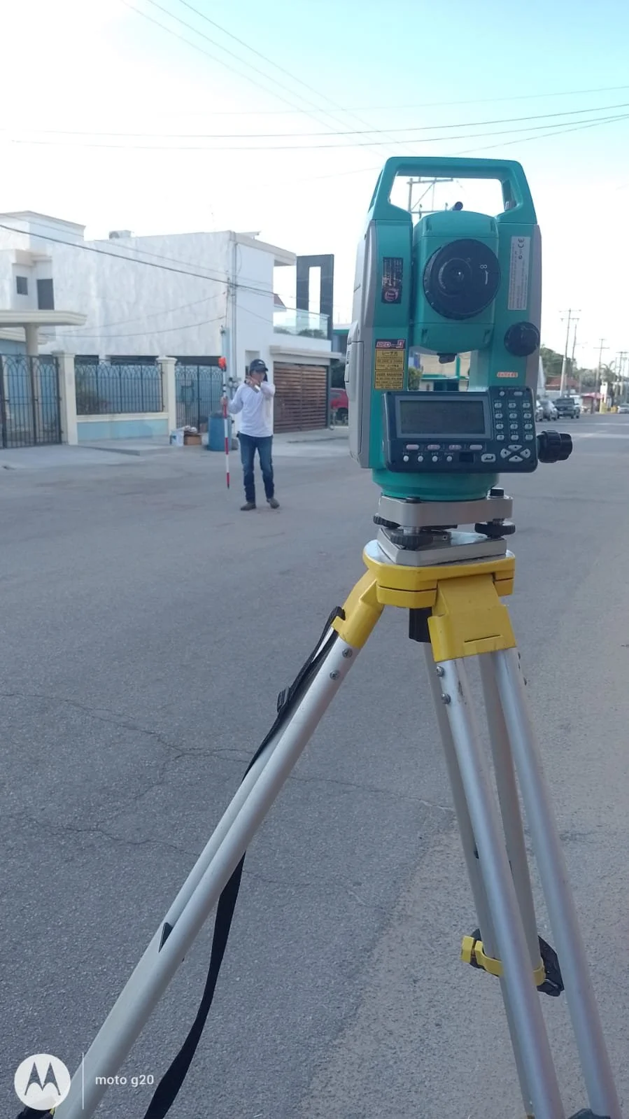 Una estación total en primer plano en una calle y una persona con equipo de medición en el fondo.