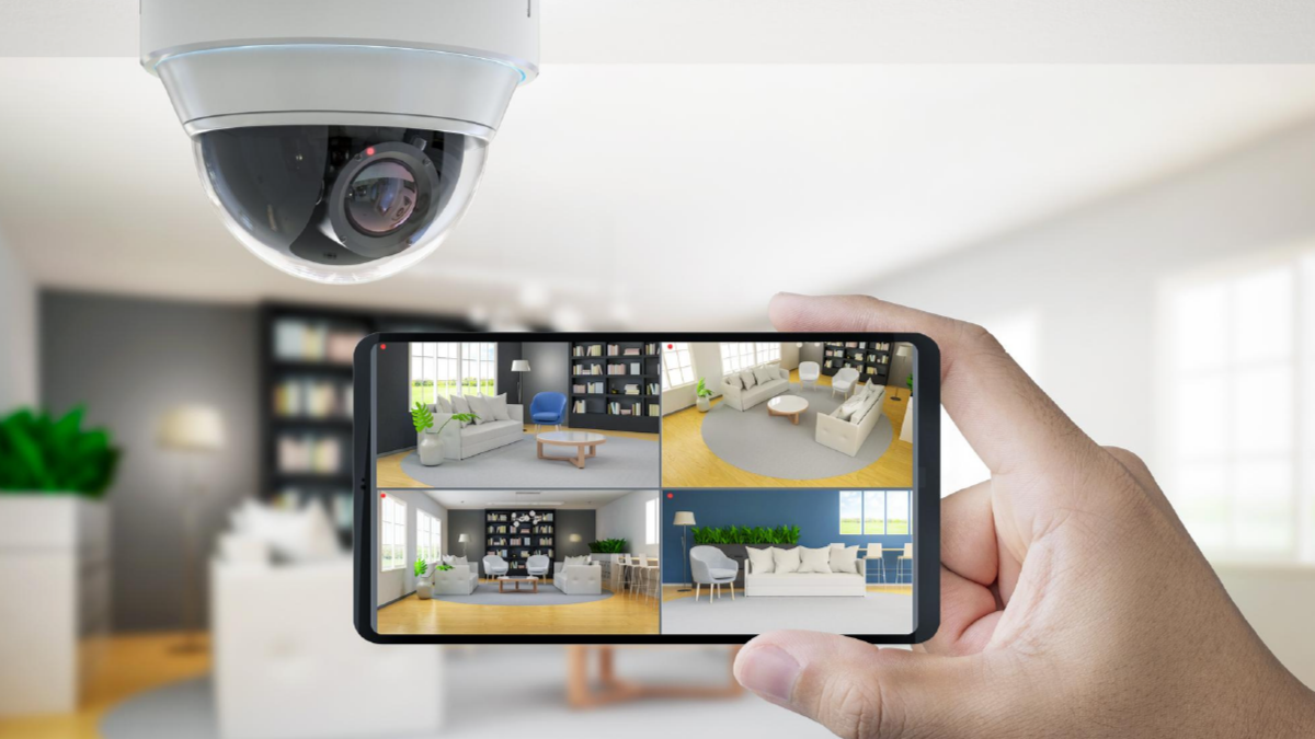 A security camera installing overhead with a person holding a smartphone displaying four different camera views of a living room interior.
