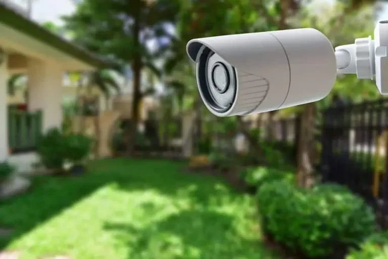 Close-up of a surveillance camera mounted outdoors overlooking a backyard with grass, trees, and a wooden fence.