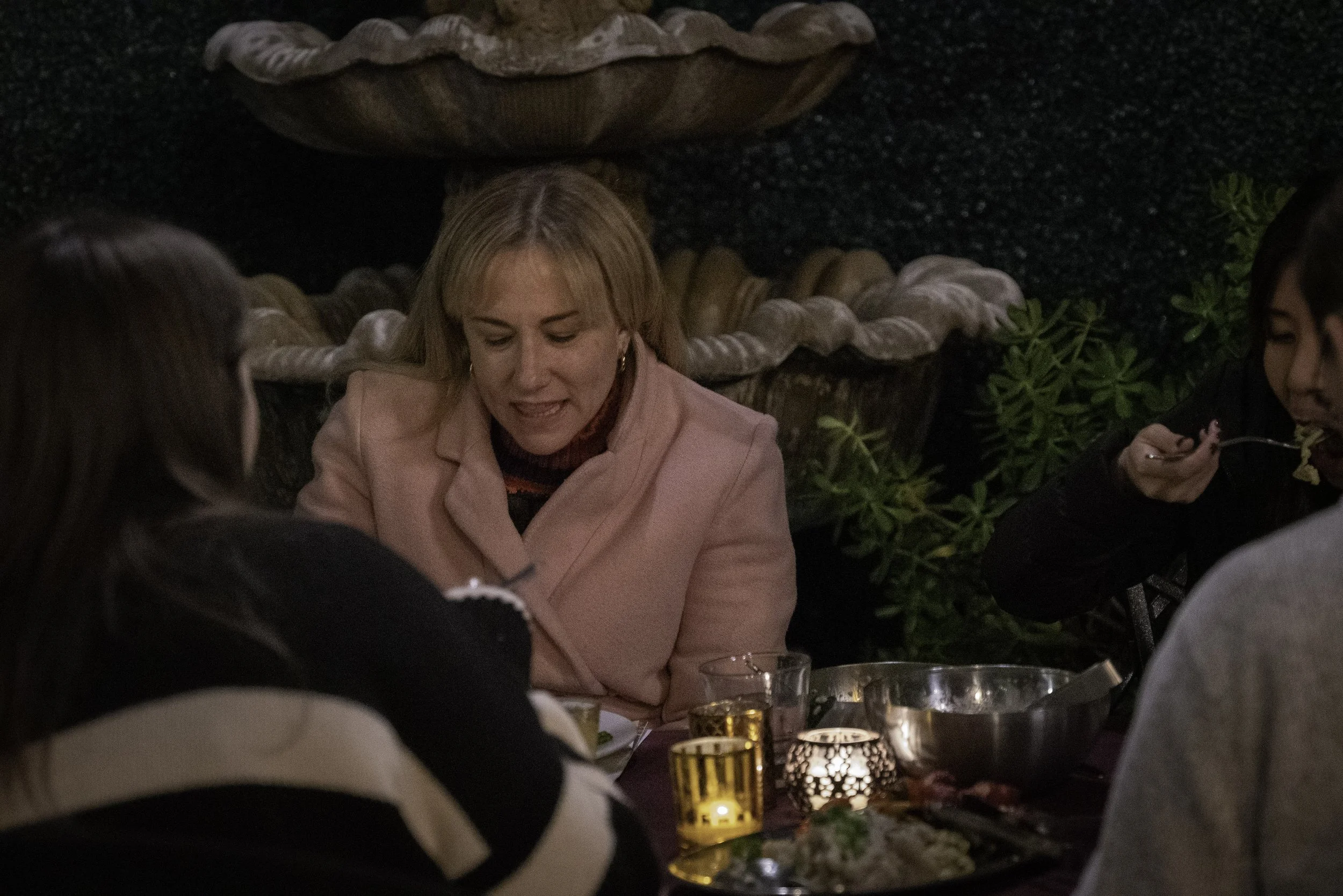  Woman in a pink coat in conversation with other attendees at a candlelit outdoor dinner event. 