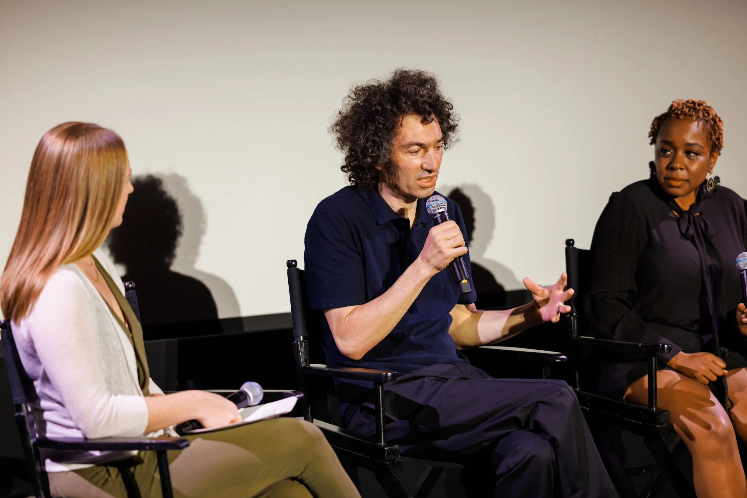  Three-person panel discussion at a film screening Q&amp;A, with a moderator and two speakers seated in director's chairs. 