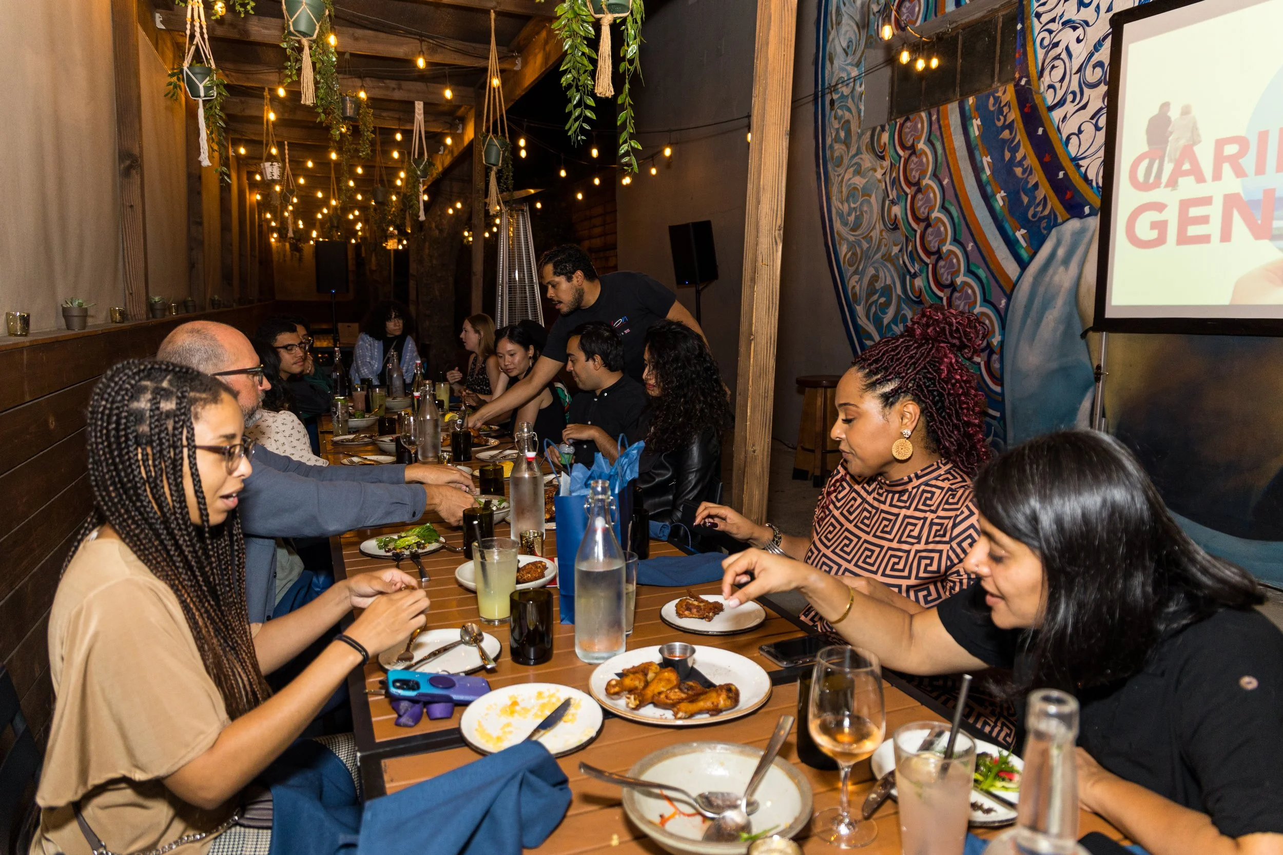  Attendees dining at a long table during a Caring Across Generations evening event, with a branded screen visible in the background. 