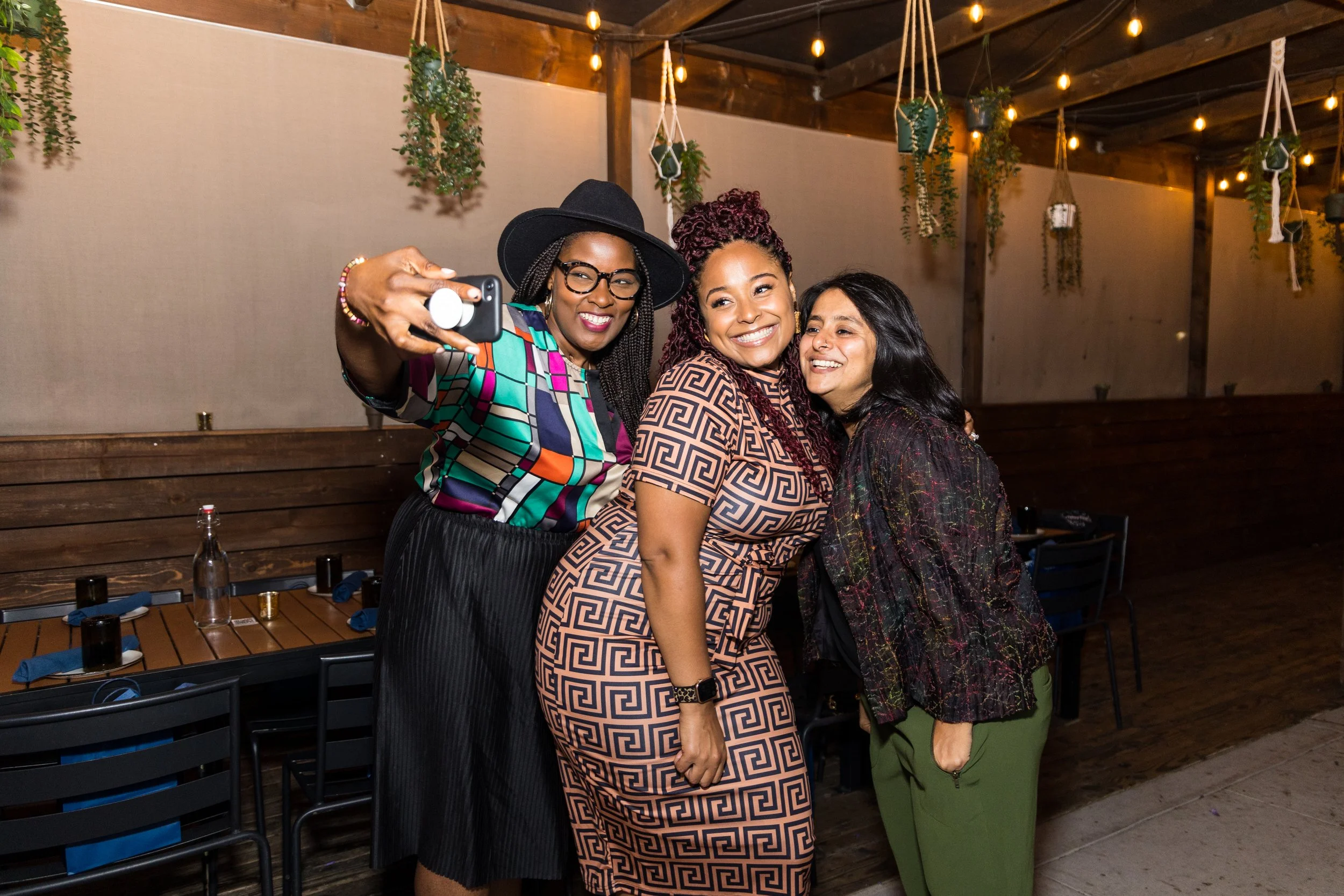  Three women smiling and taking a selfie at an indoor event space with string lights and hanging plants. 
