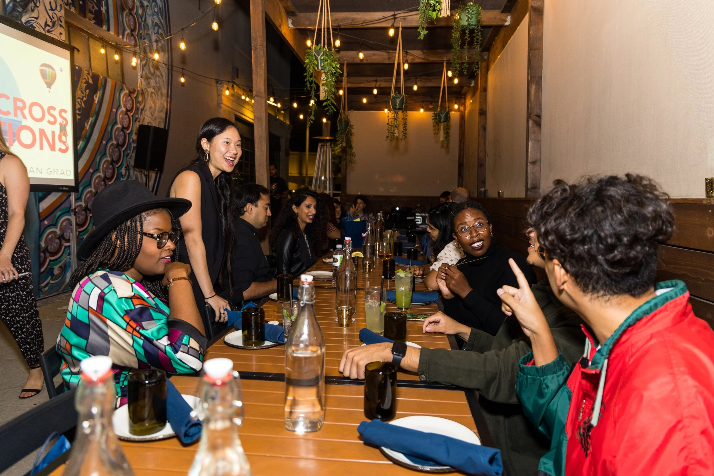  Diverse group of attendees laughing and conversing at a long dinner table during an evening event, with a Caring Across Generations presentation visible on a screen in the background. 