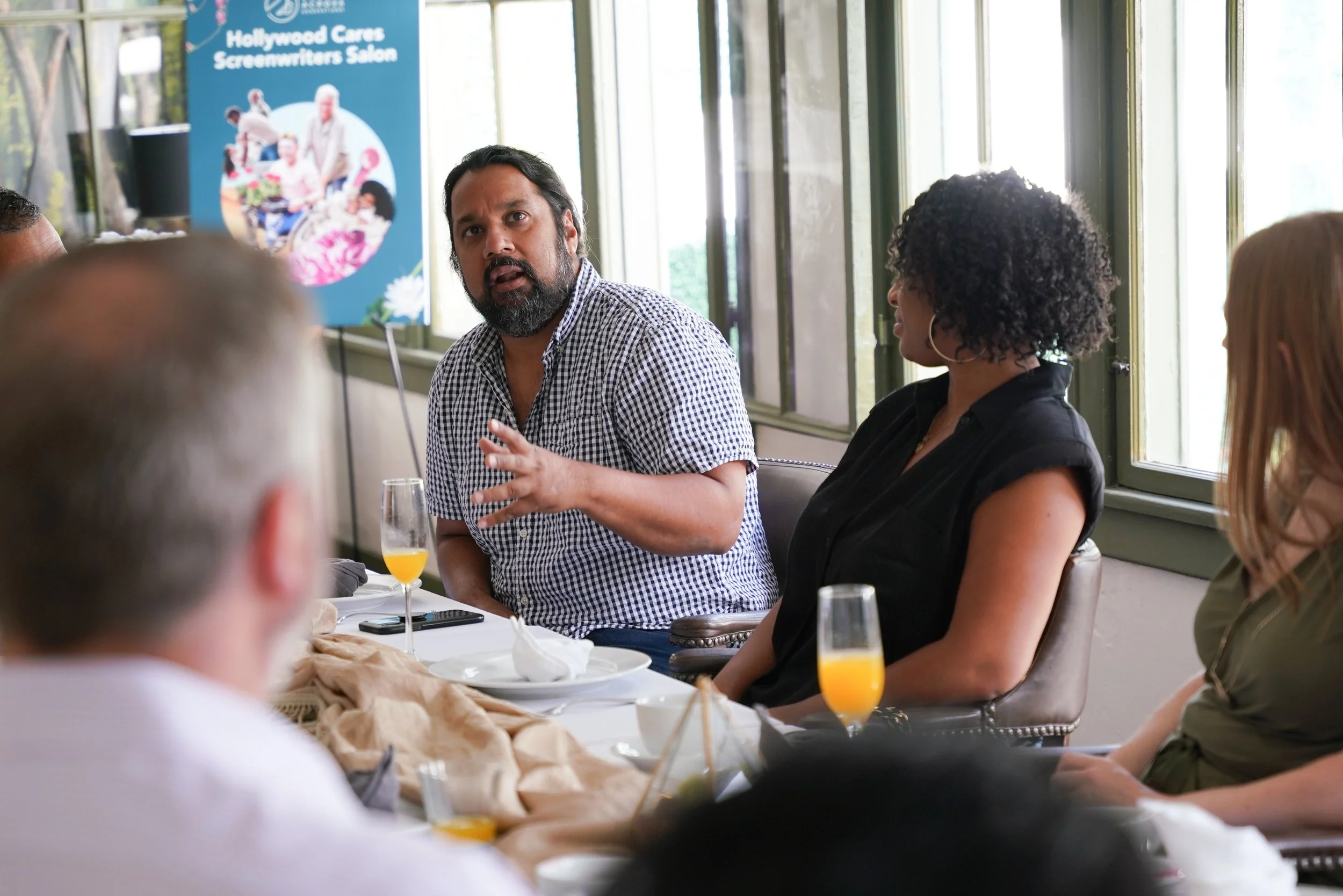  Guest speaking at the Hollywood Cares Screenwriters Salon dinner, with the event banner visible in the background. 