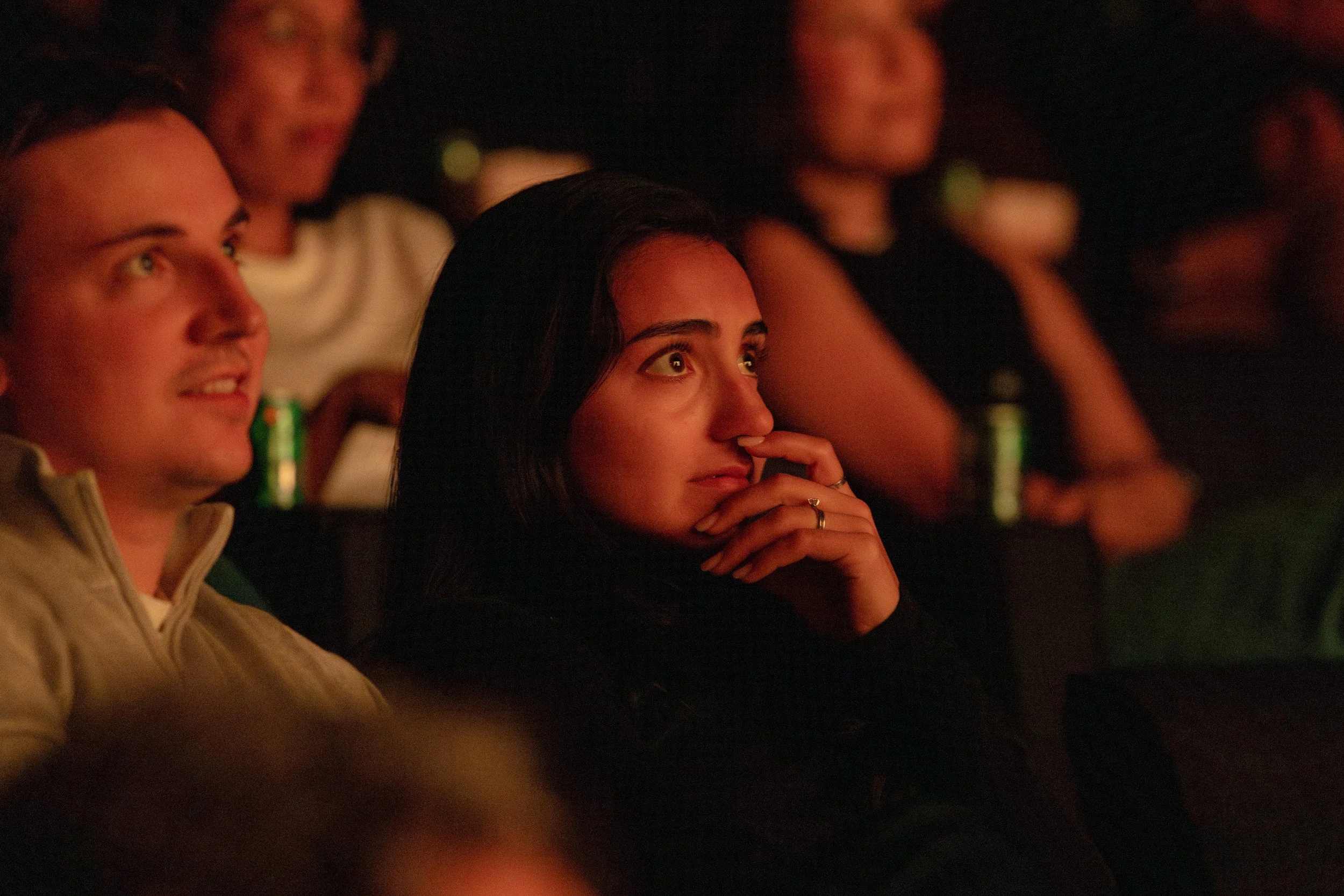  Audience members watching a film screening in a darkened theater, their faces lit by warm red light, with expressions of focused attention. 
